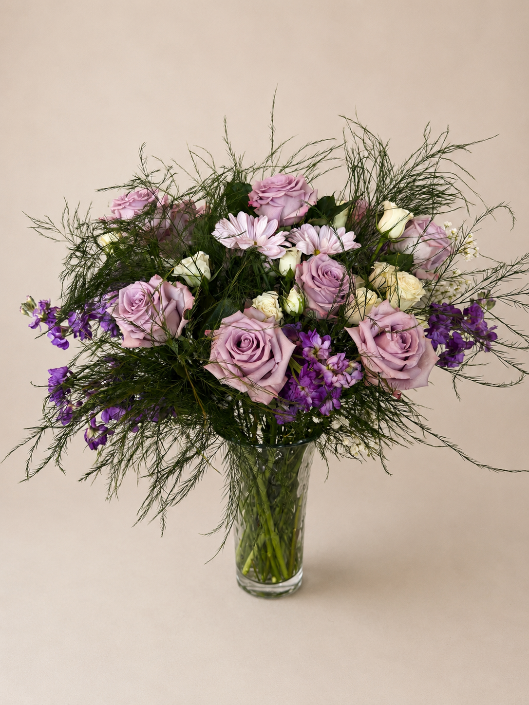 A bouquet of pink, purple, and white flowers in a clear glass vase against a neutral background.