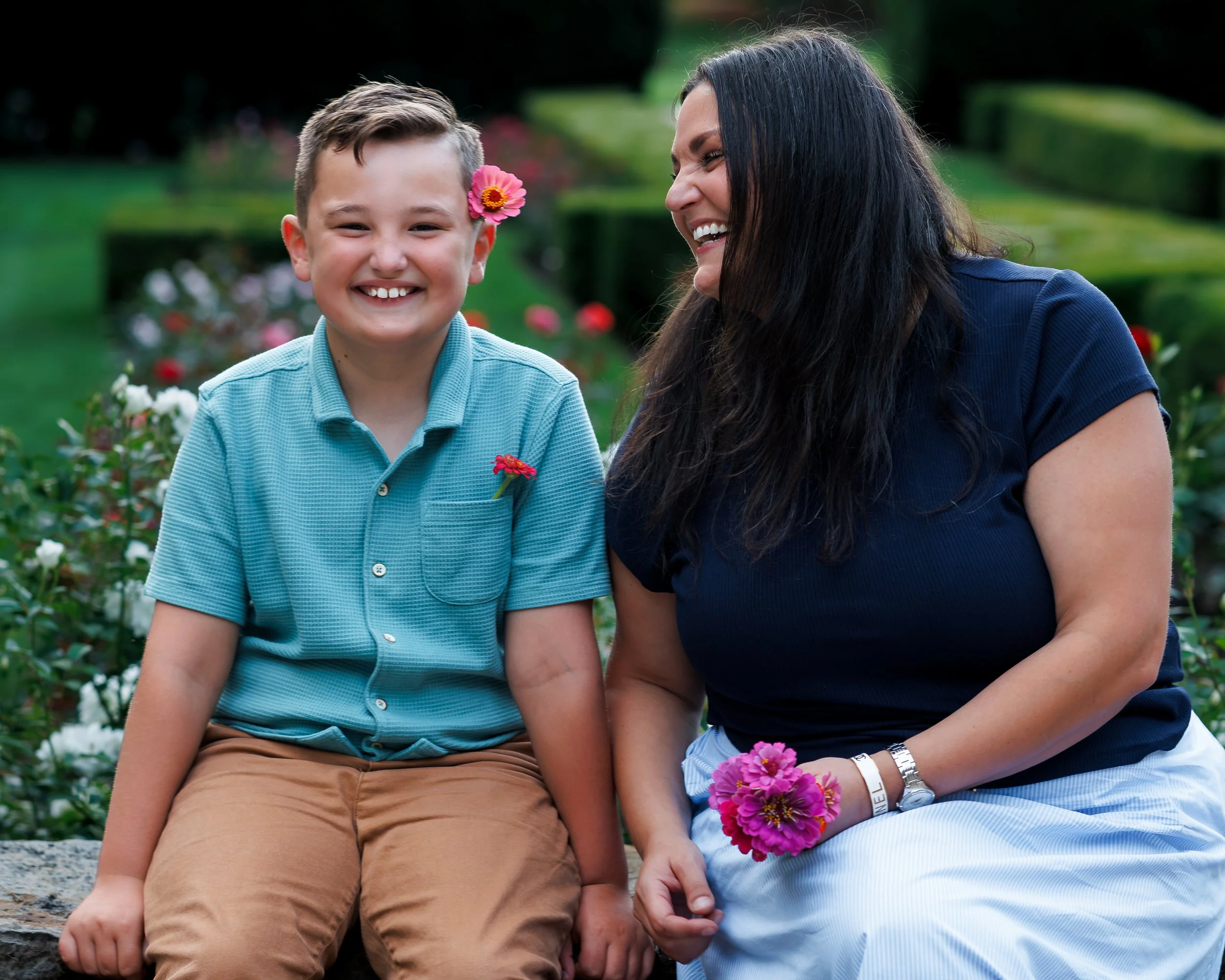 A woman and a boy sitting on a stone, smiling and sharing a moment outdoors with flowers in the background. The woman has long dark hair and is wearing a dark blue shirt, while the boy has short light brown hair, a flower in his hair, and is wearing 