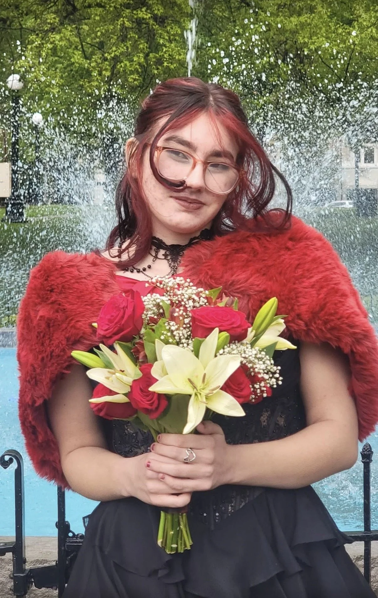 A woman with red hair and glasses holding a bouquet of red roses and white lilies, wearing a red fur stole and black dress, standing outdoors near a fountain with water splashing in the background.