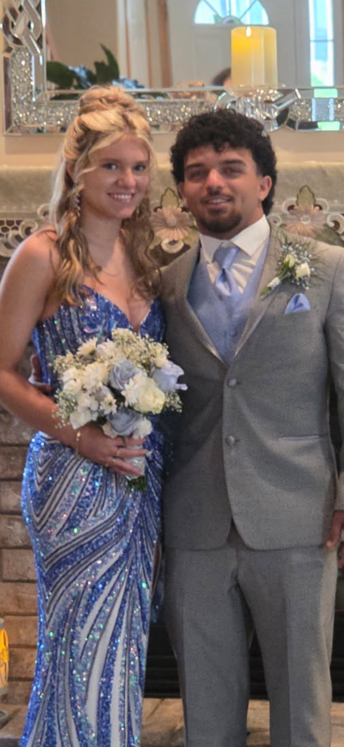 A wedding couple, woman in a blue and silver dress holding a bouquet, man in a gray suit with boutonniere, standing in a decorated venue with candle and mirror in the background.