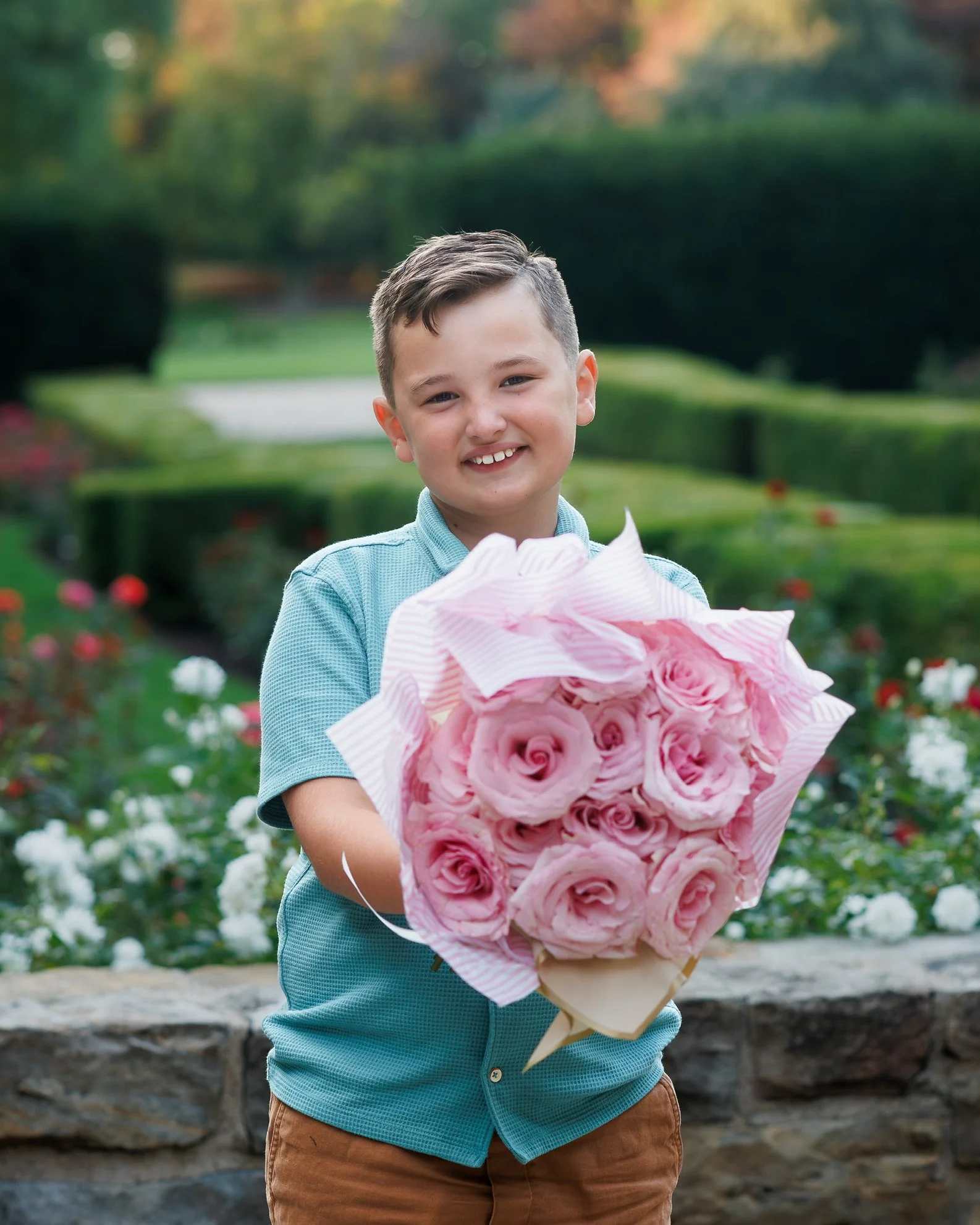 A young boy smiling and holding a bouquet of pink roses in an outdoor garden with green bushes and flowers in the background.