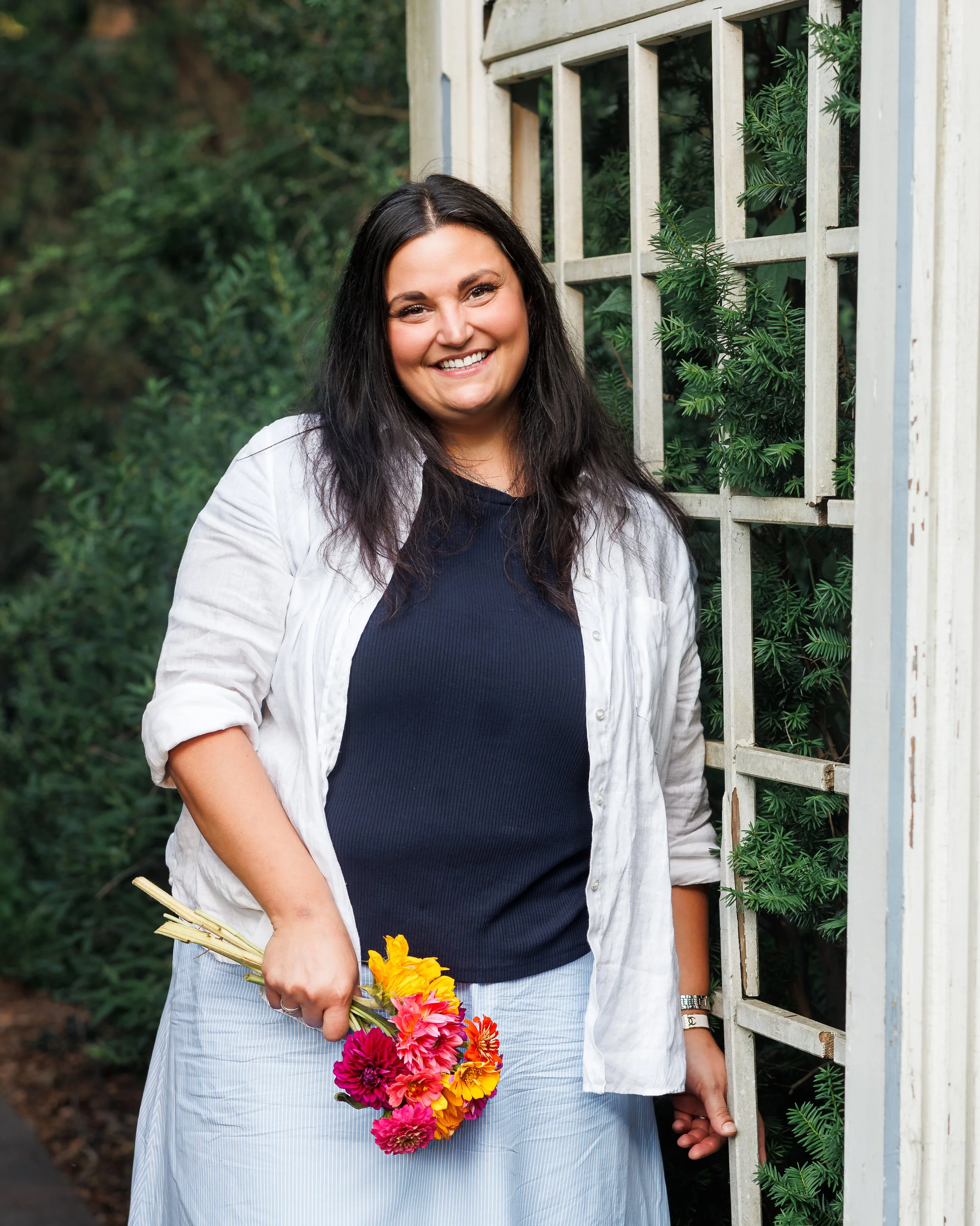 A woman with long dark hair smiling, holding a colorful bouquet of flowers, standing outdoors next to a white wooden garden fence with greenery behind her.