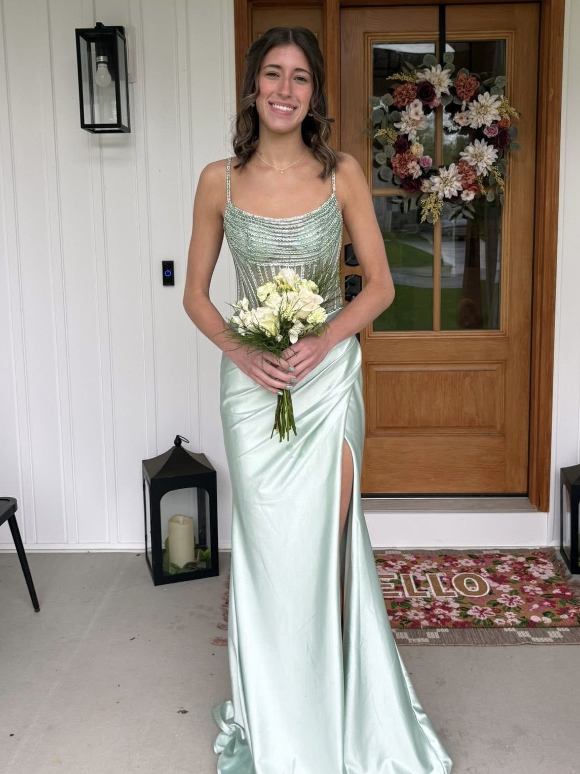 A smiling woman in an elegant light green satin dress with a slit, holding a small bouquet of white flowers, standing outside a house with a decorated door and porch.