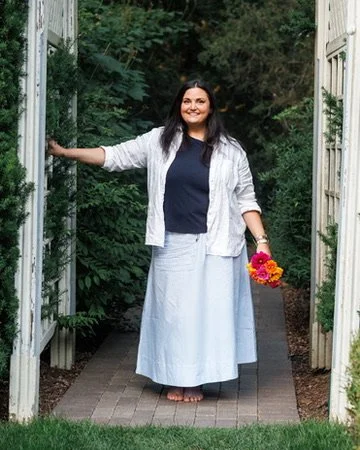 A woman standing barefoot on a brick pathway, holding a bouquet of colorful flowers, smiling, and standing in an open garden doorway with lush green plants outside.