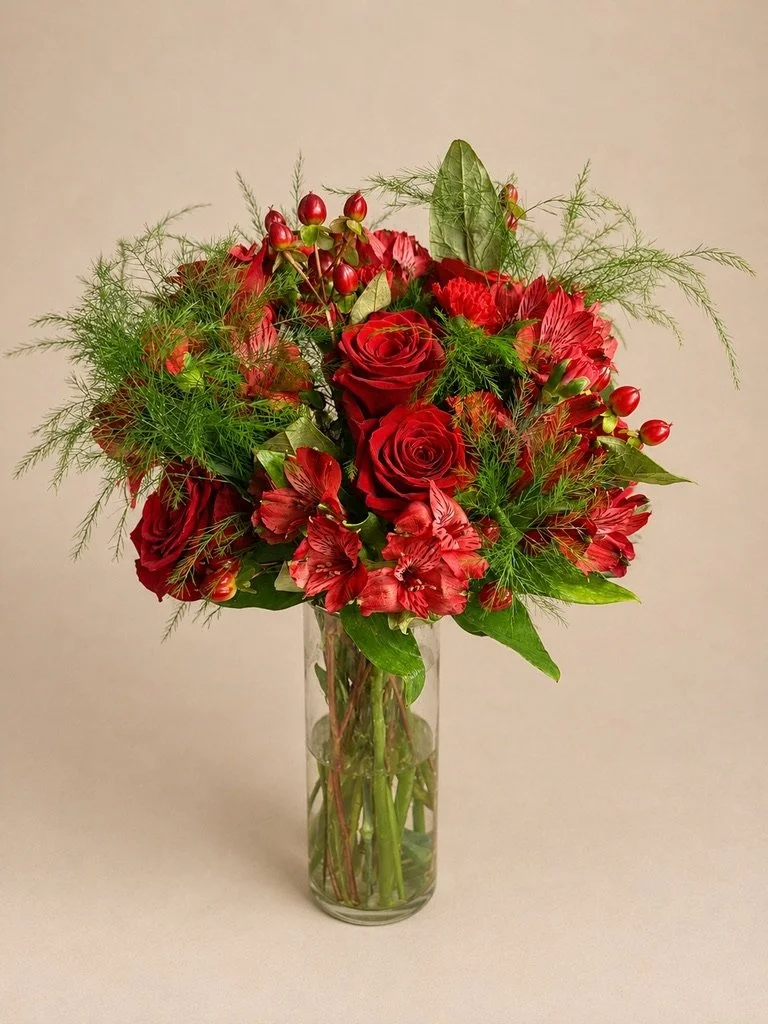 A bouquet of red roses, red alstroemeria, holly berries, and green foliage in a tall clear glass vase.