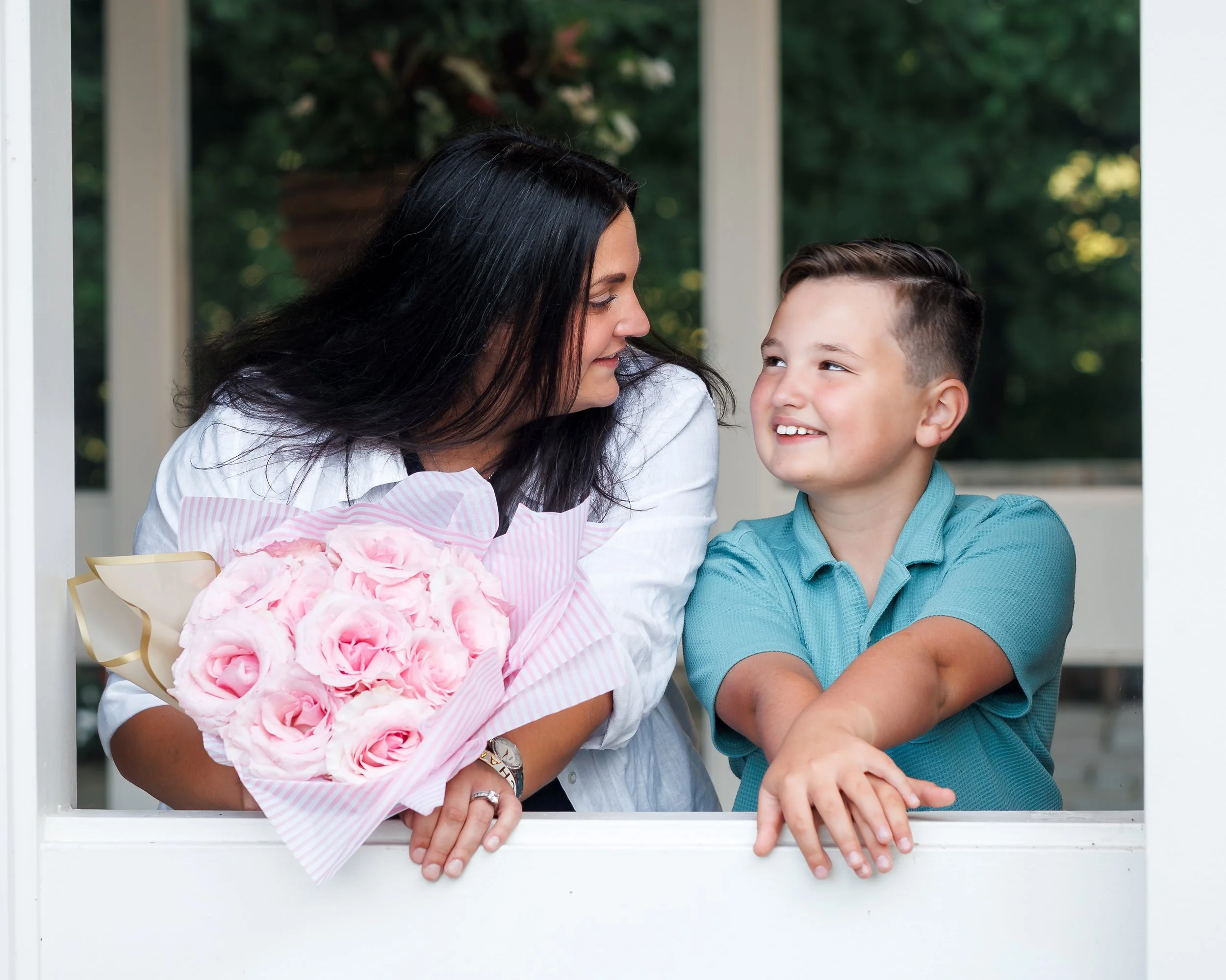 A woman and a young boy are looking at each other and smiling through a white window frame. The woman is holding a bouquet of pink flowers and has dark hair, while the boy has short brown hair and is wearing a teal shirt. Green trees are visible in t