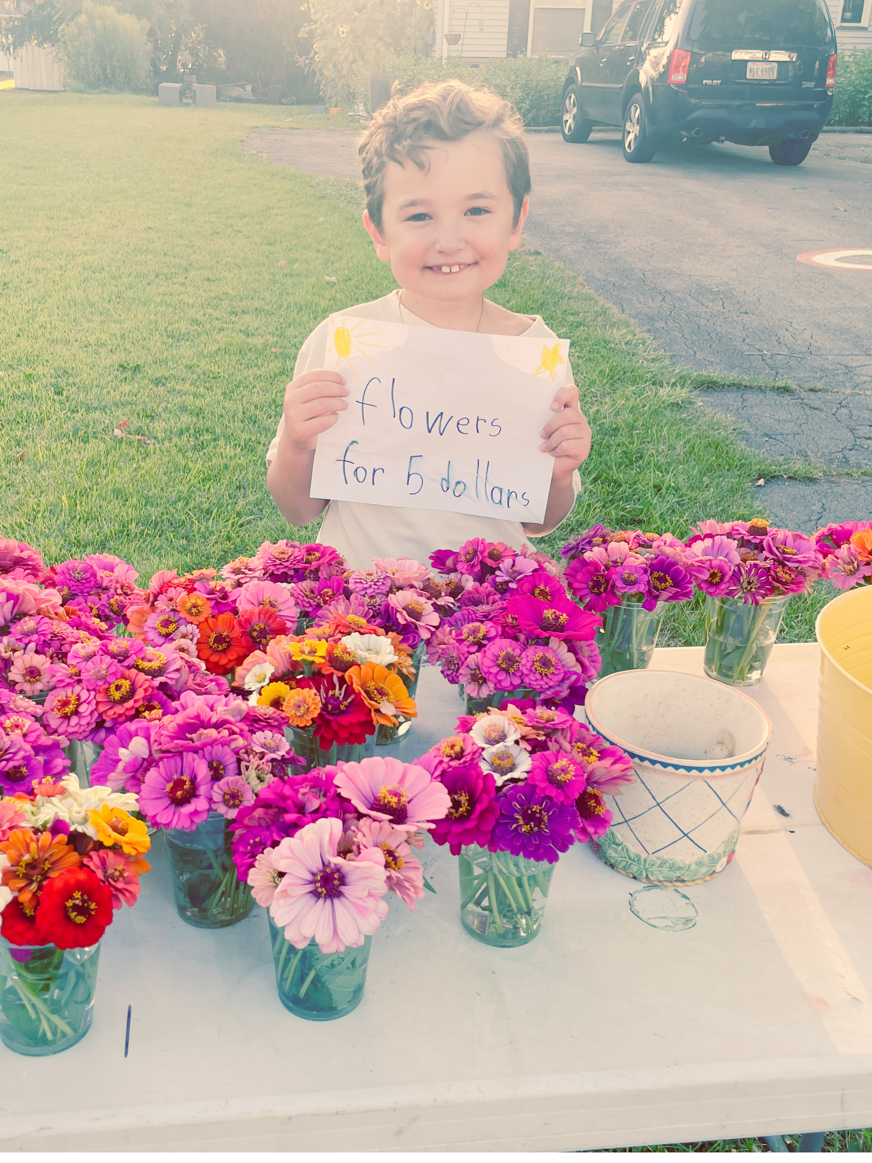 A young boy with a big smile holding a sign that says "flowers for 5 dollars" at a flower stand. The stand is filled with colorful flowers in small cups. The background shows a grassy area and parked cars.