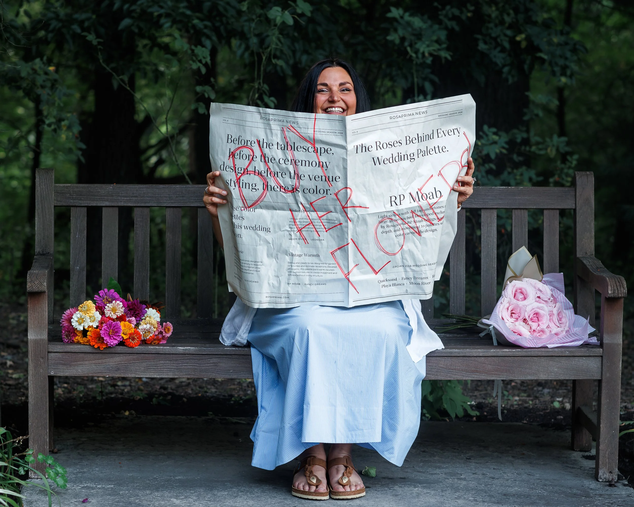 A woman sitting on a wooden bench outdoors, holding a crumpled newspaper with red handwritten text, surrounded by flowers and greenery.