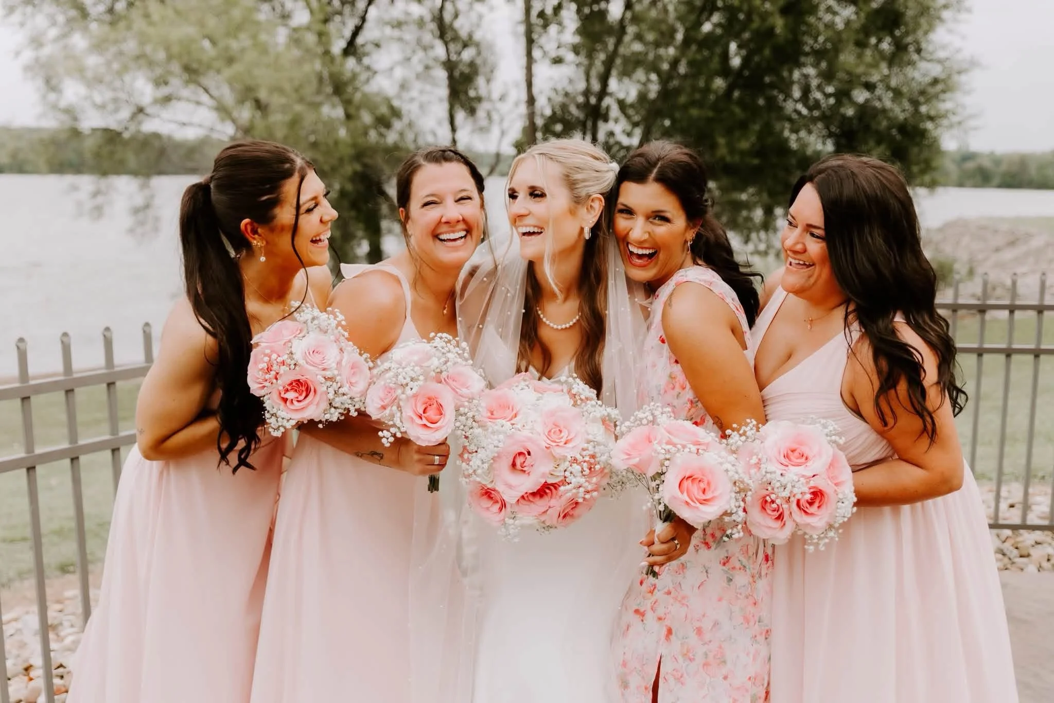 Group of five women celebrating at a wedding, holding pink and white bouquets, outdoors near a body of water and trees.