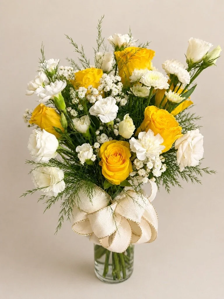 A bouquet of yellow roses, white flowers, and greenery in a glass vase tied with a cream-colored ribbon.