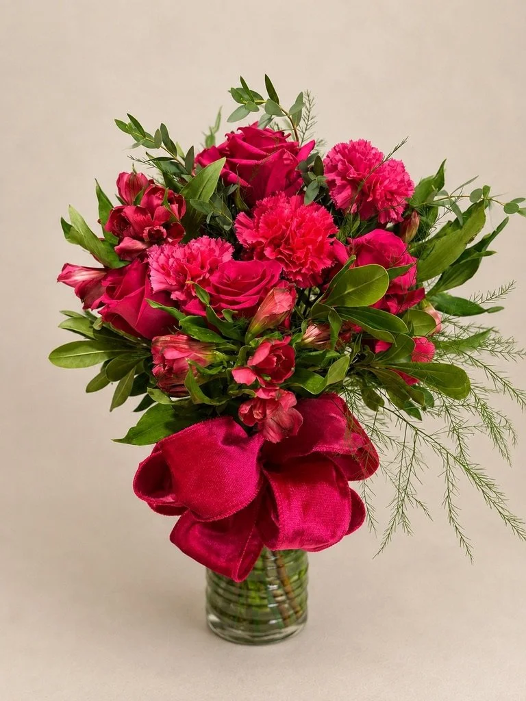 A bouquet of pink roses, carnations, and other pink flowers with green foliage in a clear glass vase, decorated with a large pink ribbon bow.