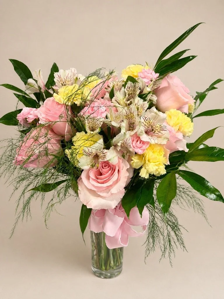A bouquet of pink, yellow, and white flowers with green leaves in a clear glass vase.