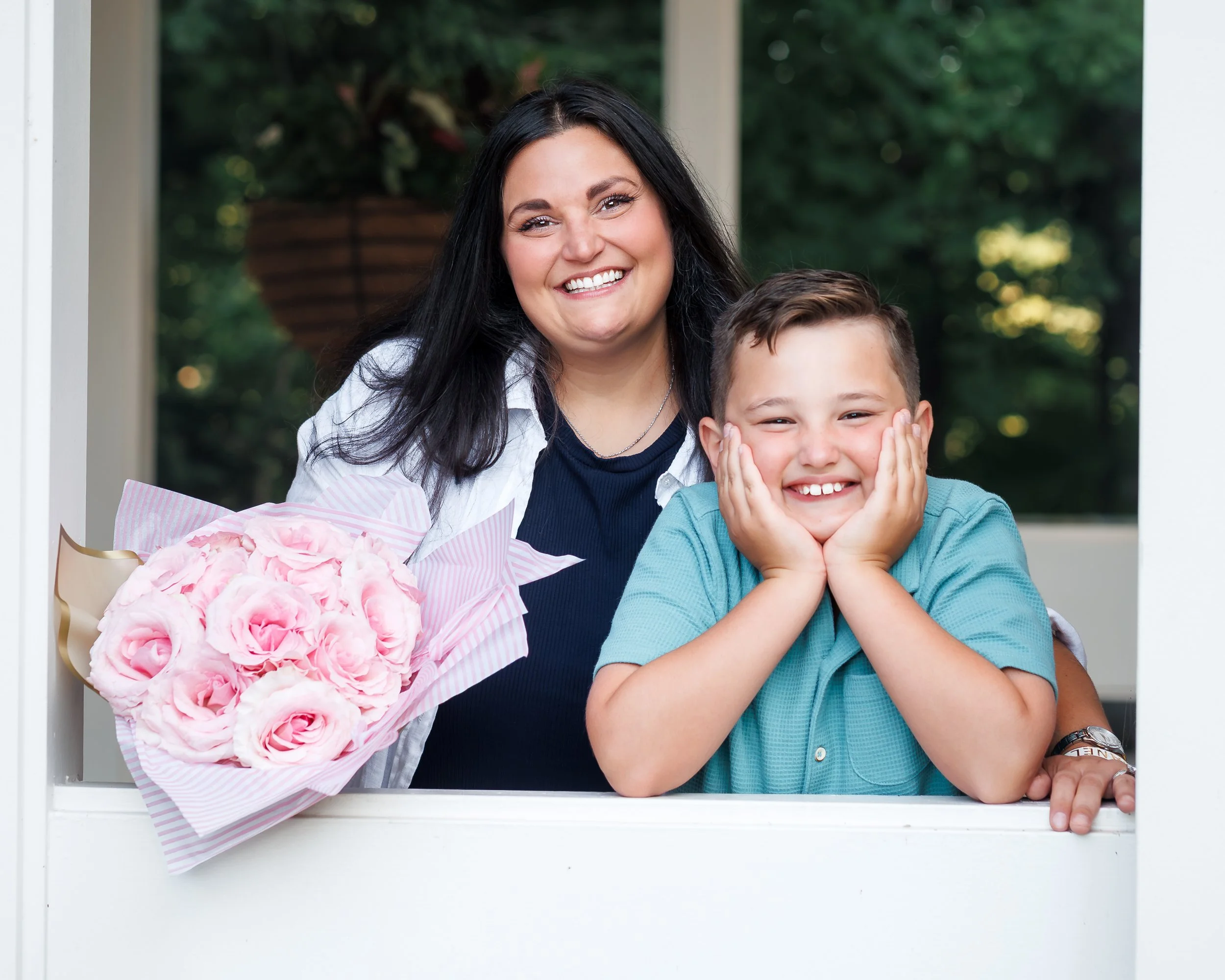 A woman with black hair and a young boy are smiling through a window. The woman is holding a bouquet of pink and white flowers. The background is green with trees.