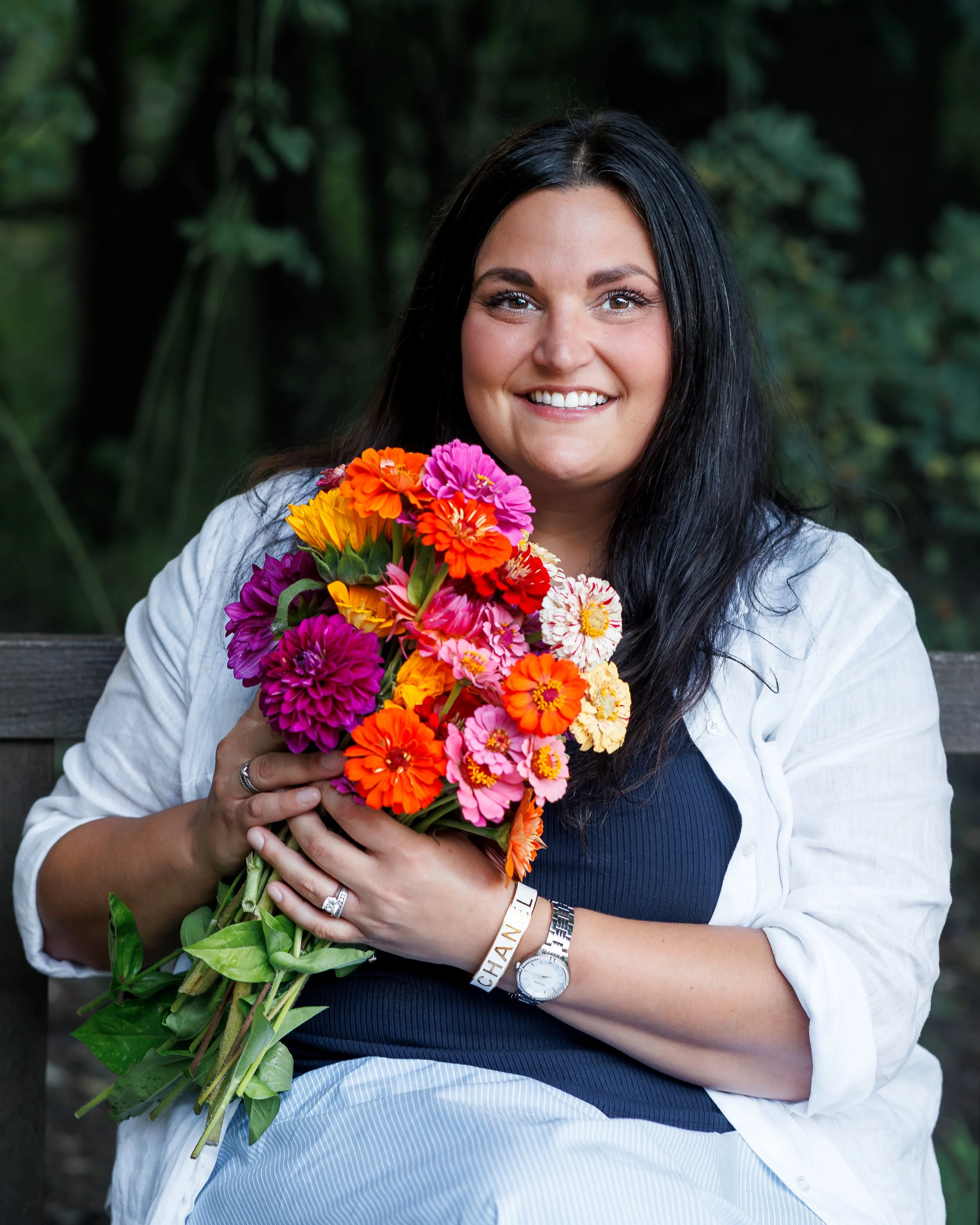 A woman with long dark hair, wearing a white shirt and a blue dress, smiling, holds a large colorful bouquet of flowers in front of her. She has a watch and bracelets on her wrist, and is seated outdoors with blurred green foliage in the background.