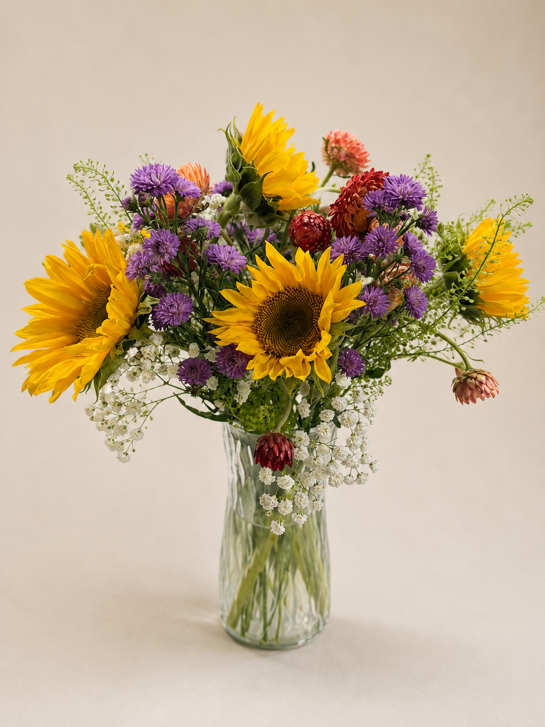 A bouquet of sunflowers, purple asters, small pink and white flowers, and greenery in a clear glass vase.