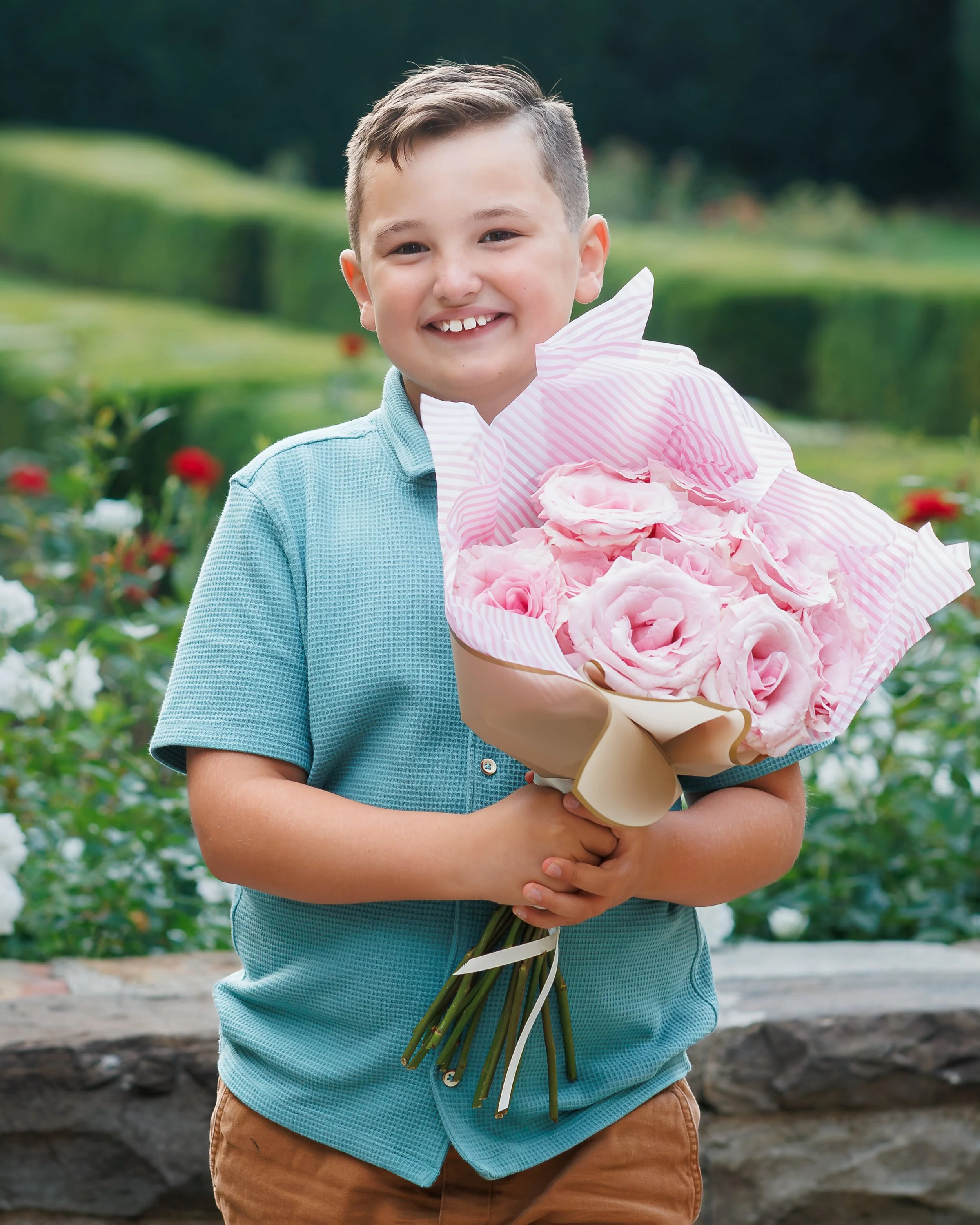 A young boy smiling and holding a bouquet of pink roses outdoors in a garden with green bushes and flowers in the background.
