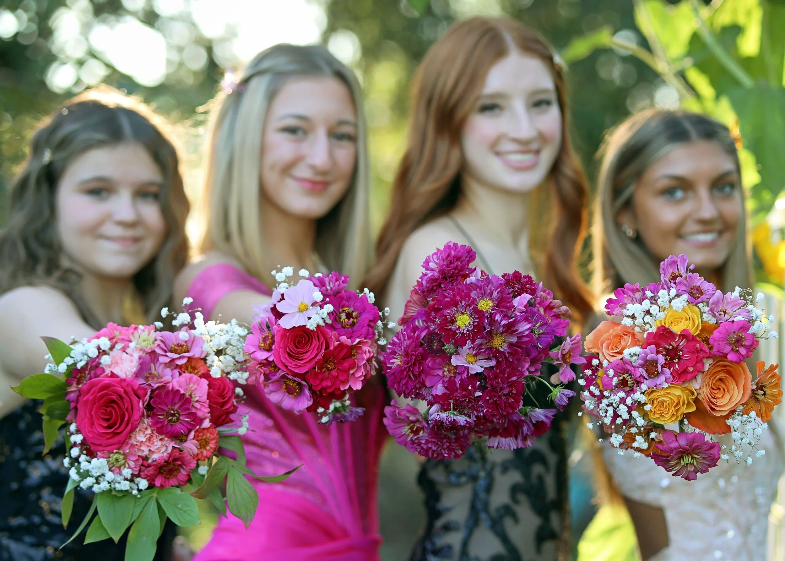 A group of women holding colorful bouquets of flowers outdoors with sunlight filtering through trees.