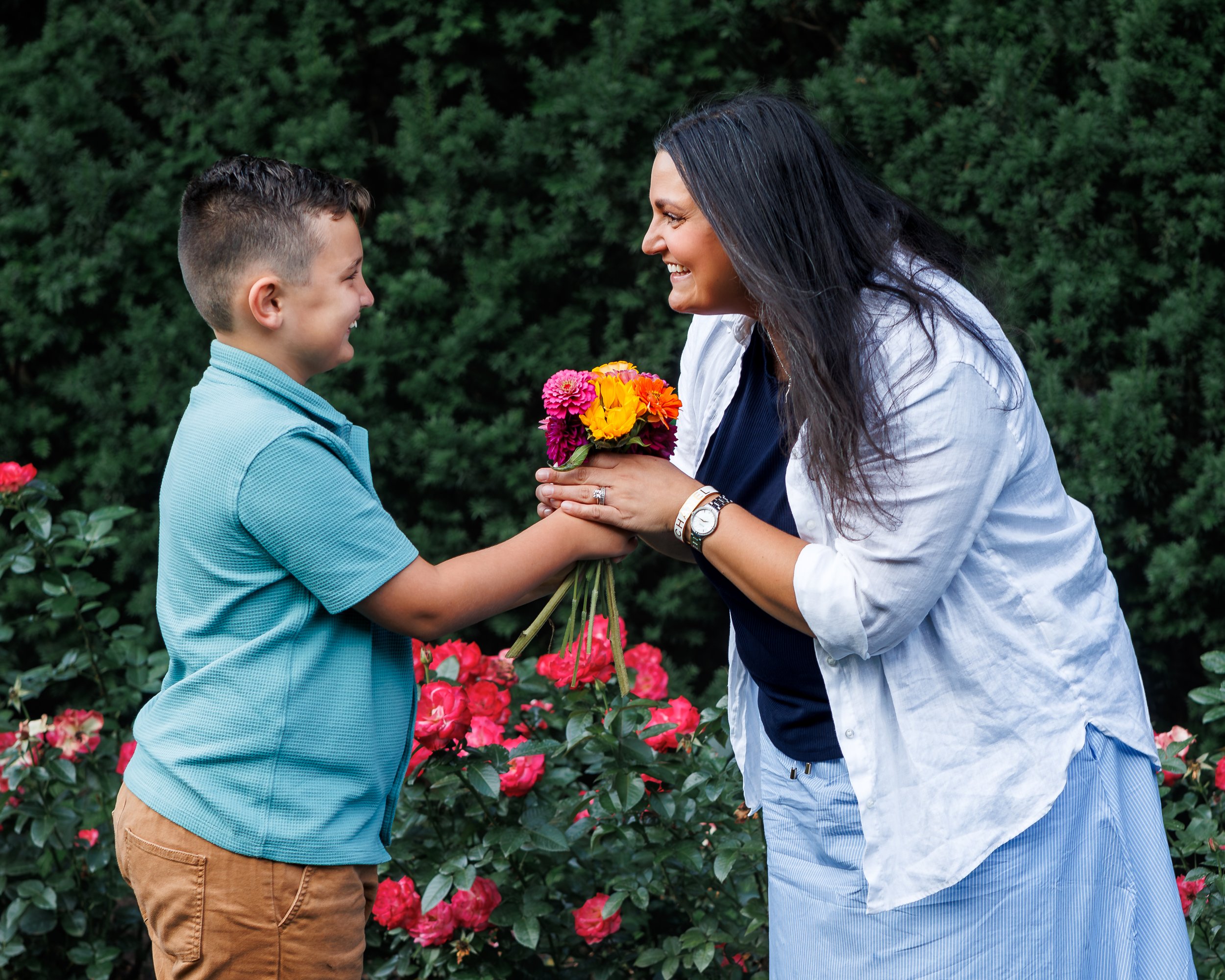 A woman and a boy face each other smiling, with the woman holding a small bouquet of colorful flowers, standing outdoors in front of green bushes and pink flowers.