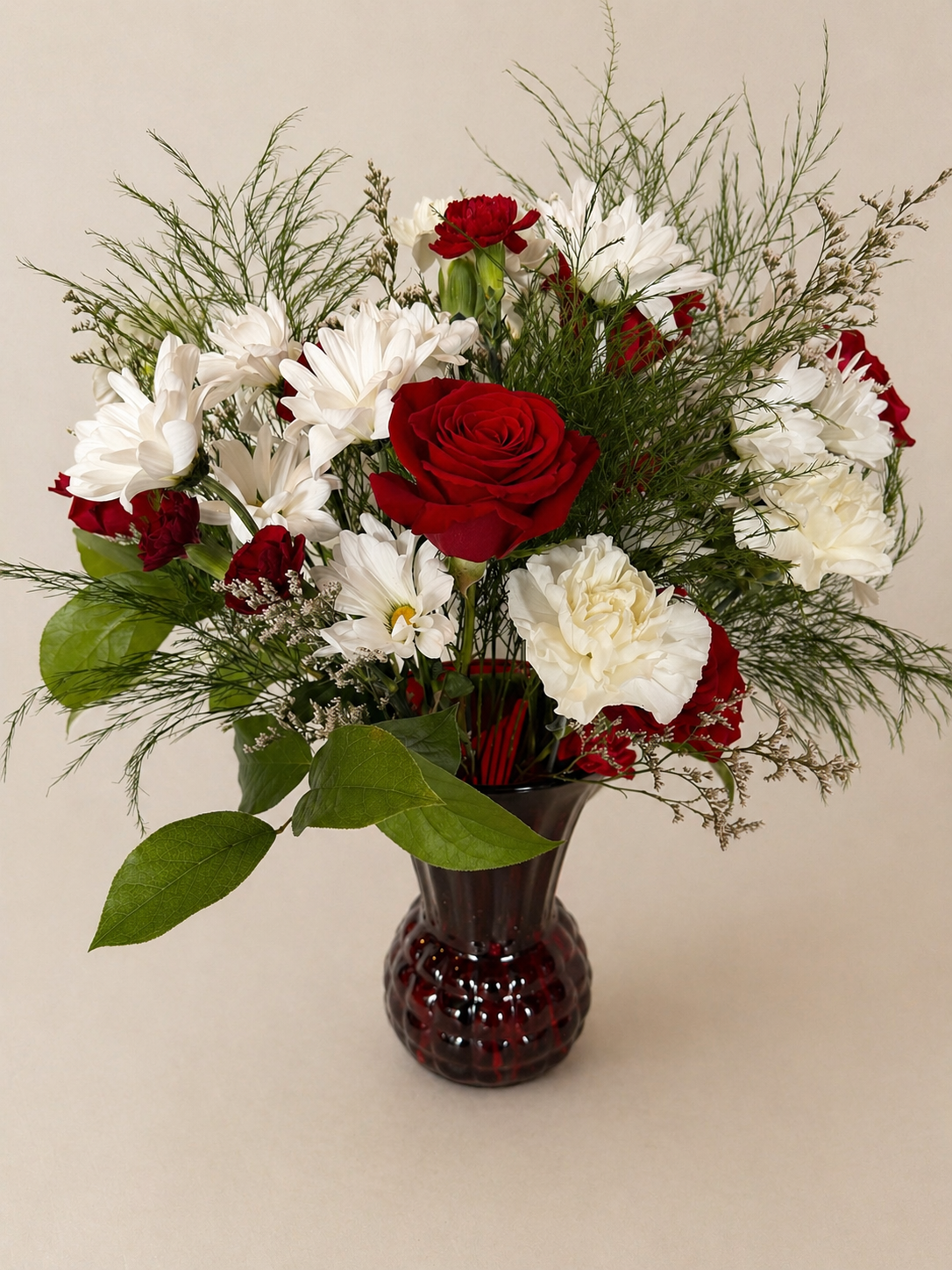A bouquet of red roses, white daisies, and white carnations in a dark red glass vase.