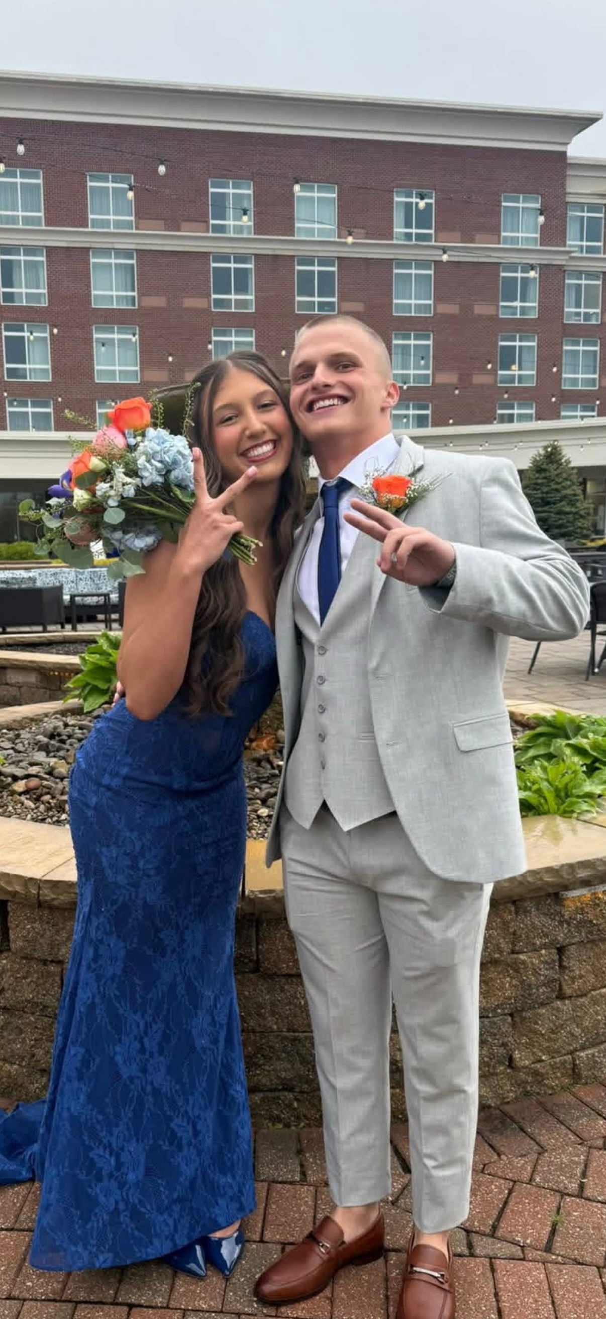 A young couple dressed in formal attire, smiling and posing together outdoors, with the woman holding a bouquet of colorful flowers. The woman is wearing a long blue lace dress and has long wavy hair, while the man is wearing a light gray suit with a