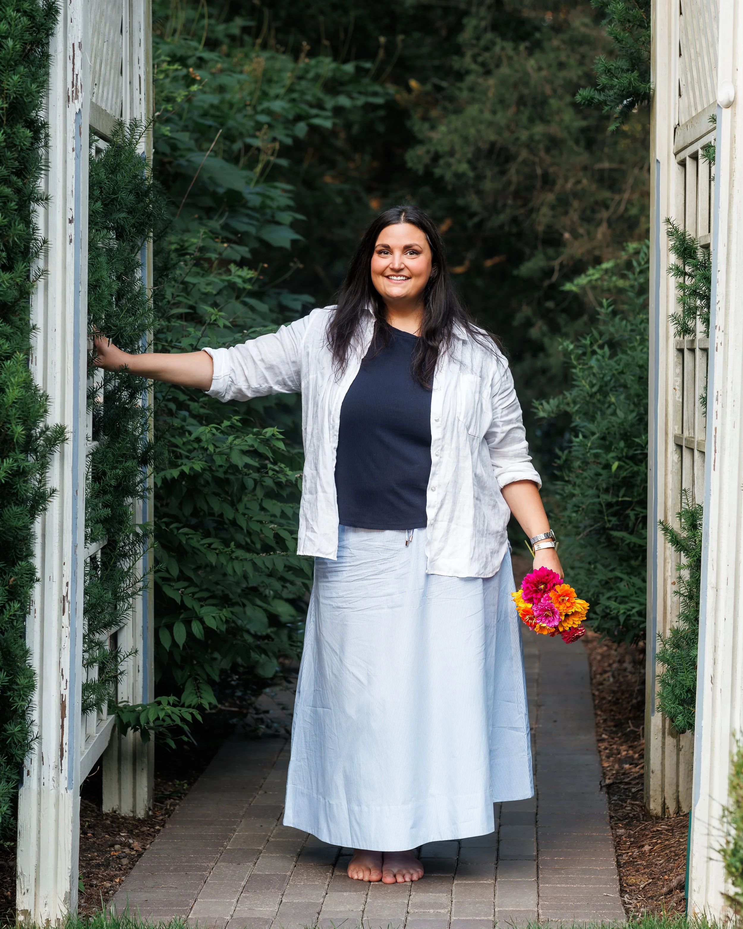 Woman smiling, standing barefoot on a brick pathway under a garden archway, holding a bouquet of colorful flowers, surrounded by green foliage.