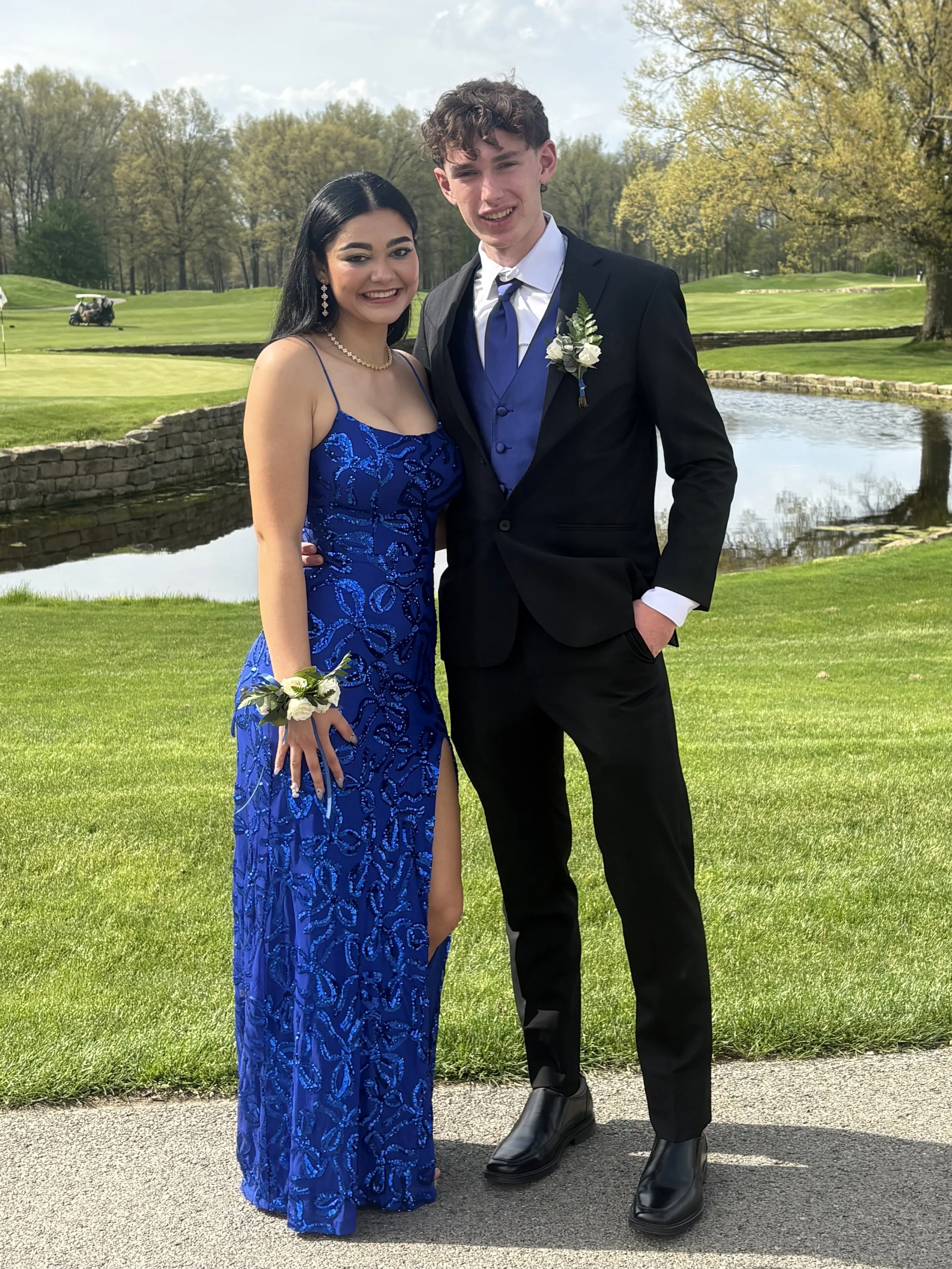 A young man and woman dressed in formal attire standing on a sidewalk near a golf course. The woman is wearing a blue evening gown with floral embroidery, a pearl necklace, earrings, and a white corsage on her wrist. The man is wearing a black suit w