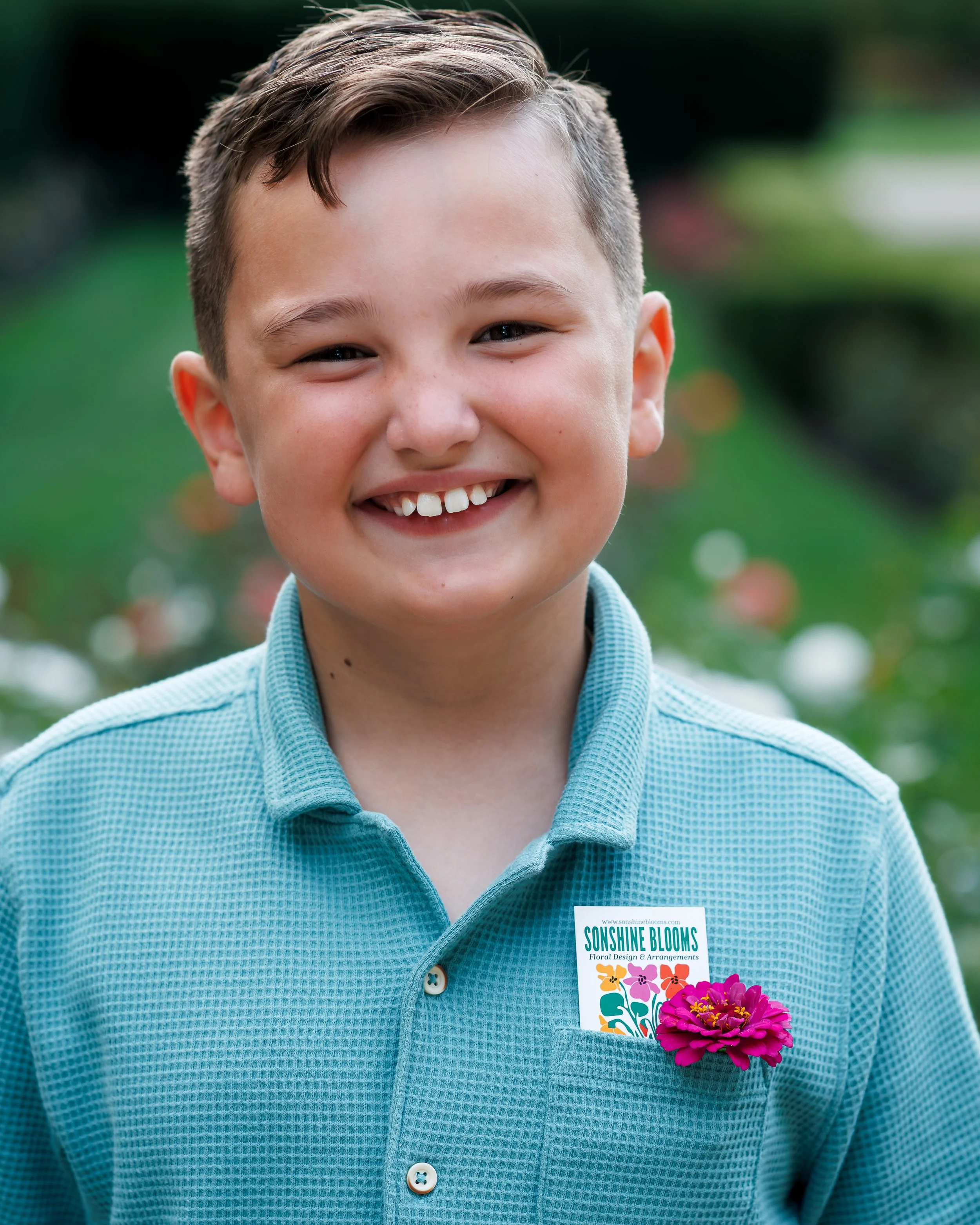 A young boy smiling outdoors wearing a turquoise button-up shirt. He has a flower bouquet in his shirt pocket and a badge that reads "Sunshine Blooms Flower Design & Arrangements." The background is green and blurred.
