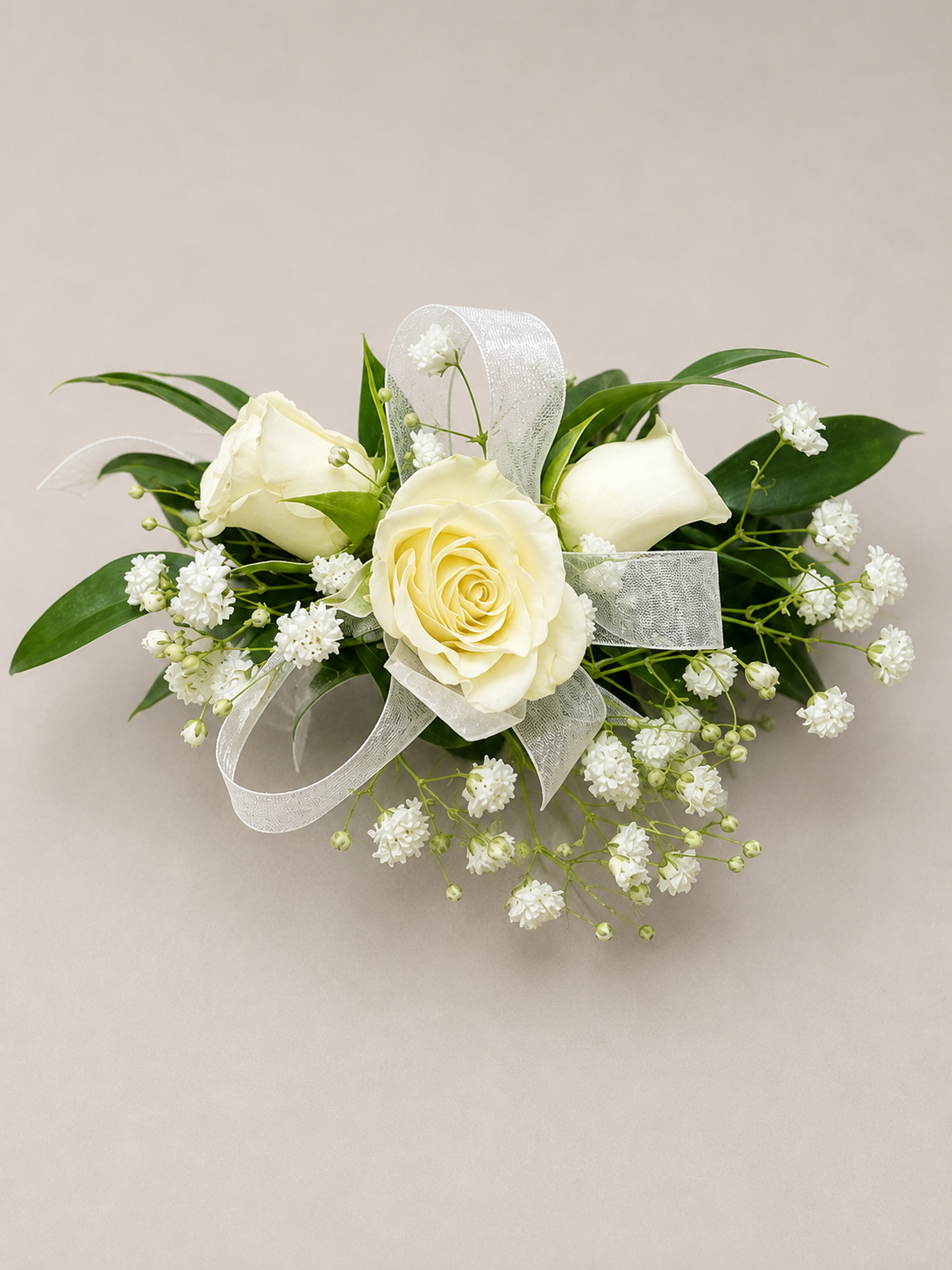 A white floral corsage with roses, bellflowers, green leaves, and a white ribbon bow, arranged on a light beige background.