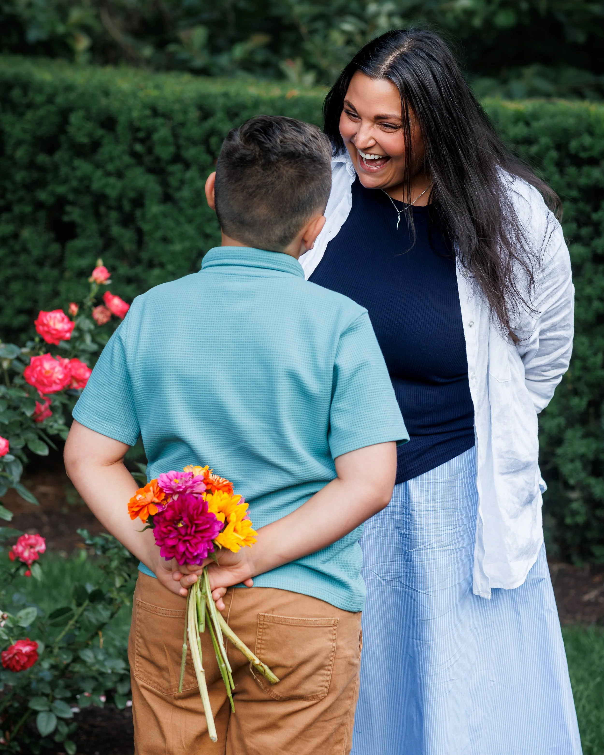 A woman with long dark hair smiling and laughing as she looks at a young boy, who is holding a bouquet of colorful flowers behind his back, in a lush green garden with pink flowers.