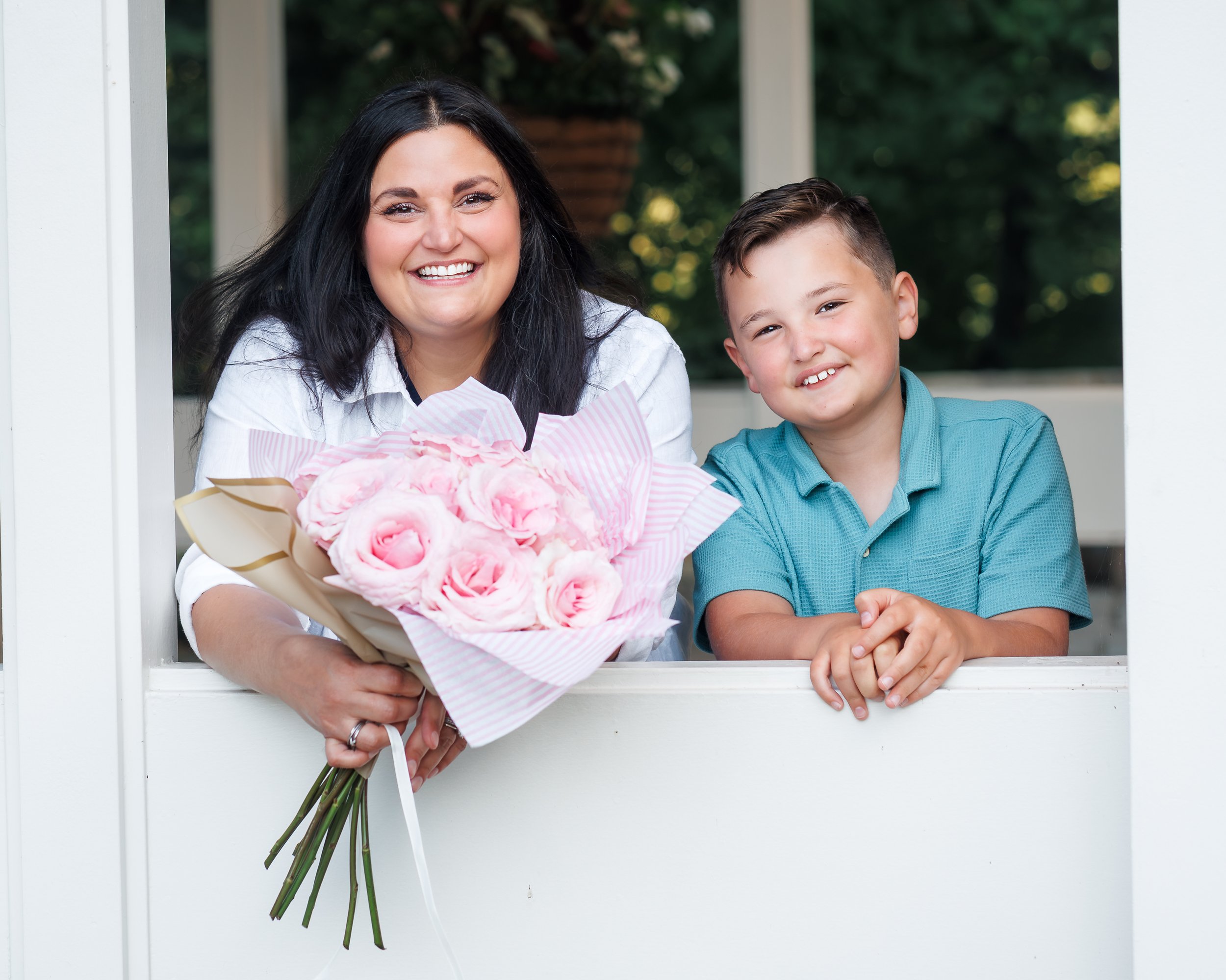 A woman with black hair and a boy with short brown hair smiling at a camera while leaning on a white porch railing, holding a bouquet of pink flowers wrapped in striped paper