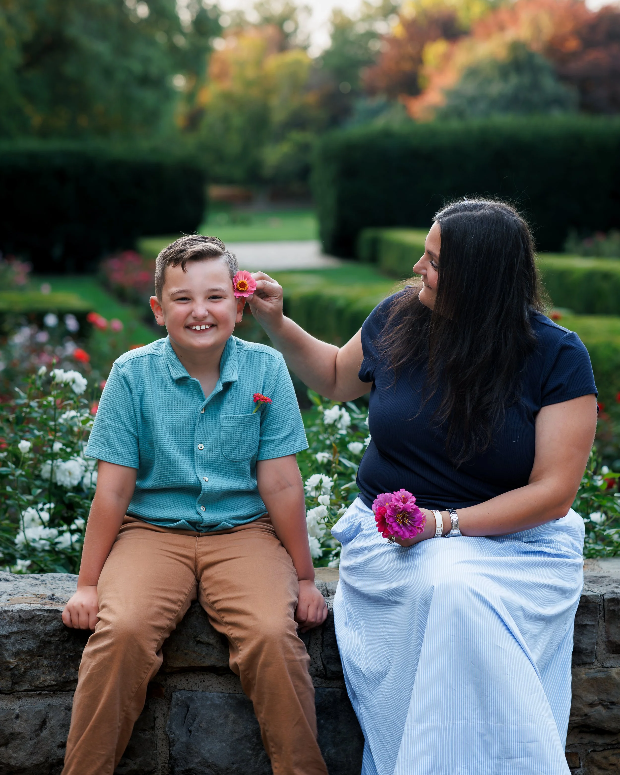 A woman and a young boy sitting outdoors in a garden, with the woman holding a pink flower near the boy's face, both smiling.