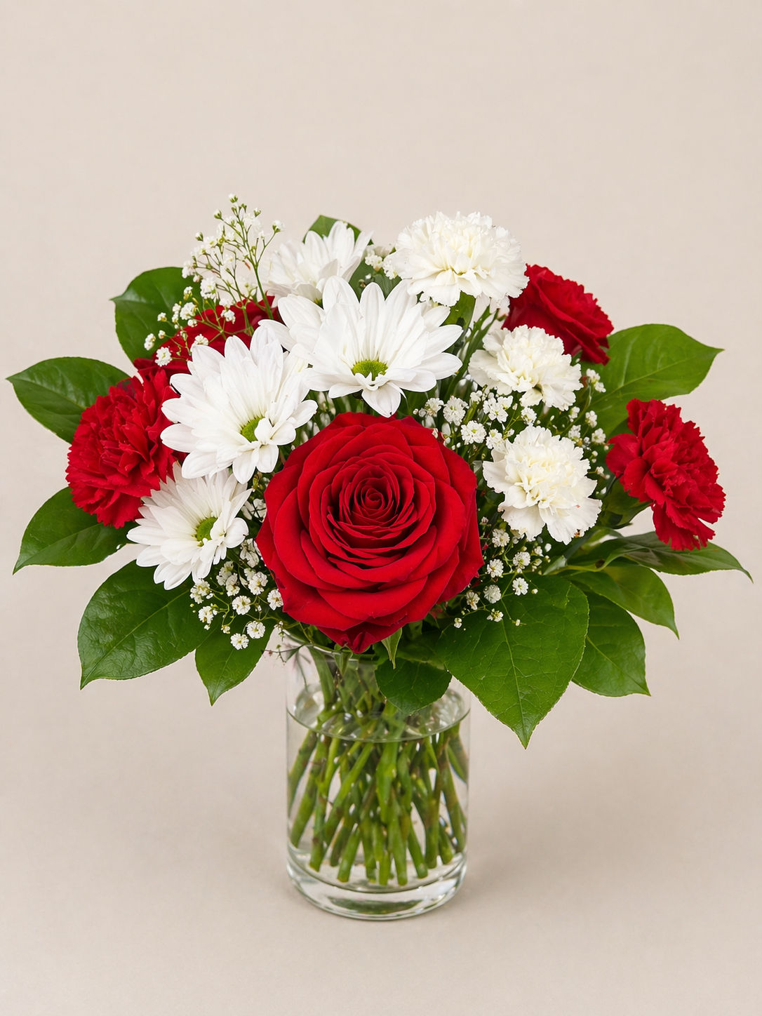 A bouquet of red roses, white daisies, white carnations, red carnations, and baby's breath in a clear glass vase with green leaves.