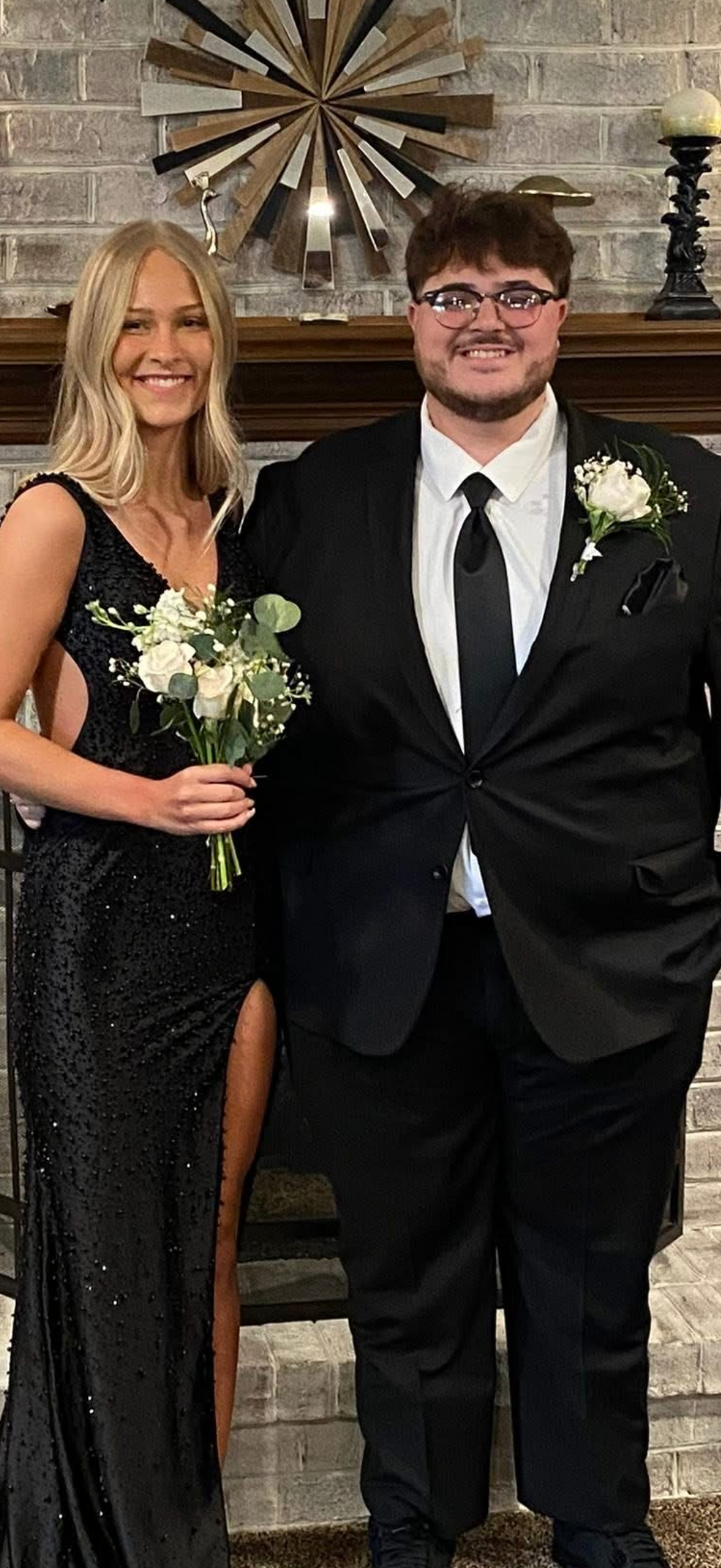 A couple dressed in formal attire, standing together in front of a brick fireplace with a modern starburst wall decoration above. The woman is wearing a long black sequined dress with a slit, holding a bouquet of white flowers, and smiling. The man i