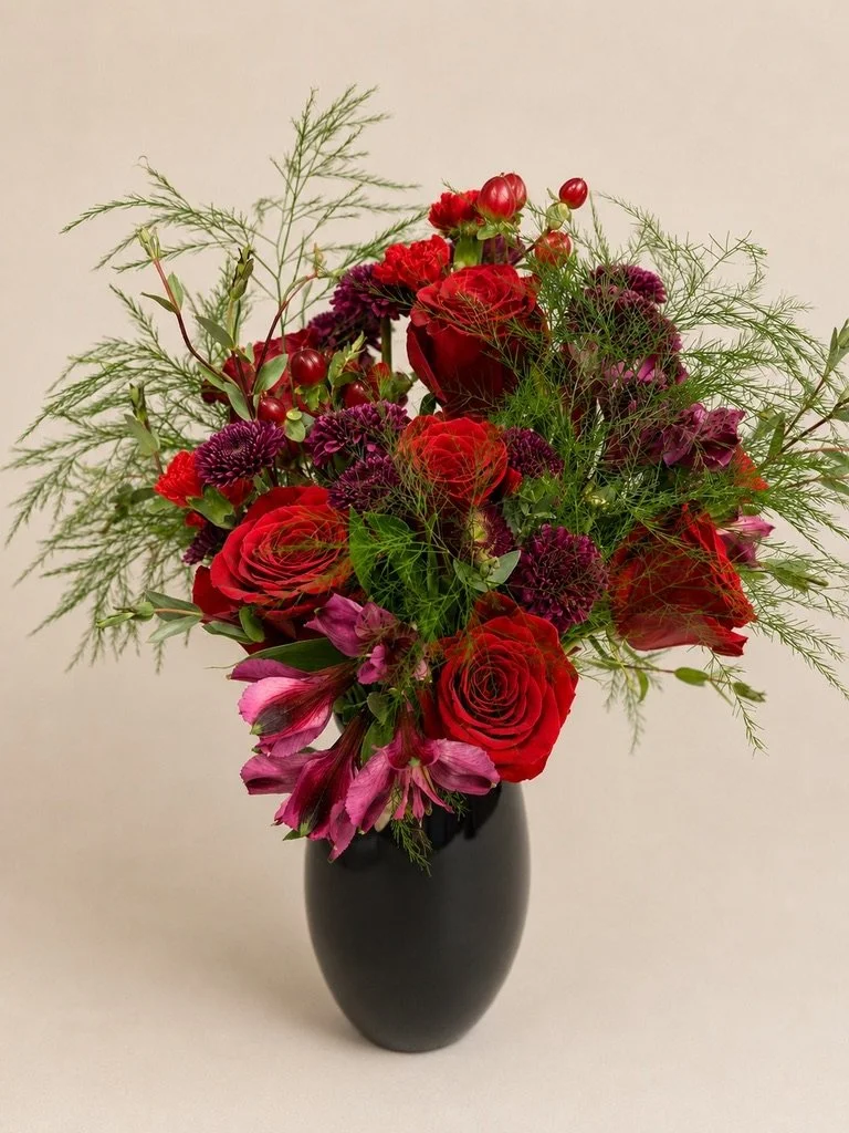 A floral arrangement in a black vase featuring red roses, purple and pink flowers, red berries, and green foliage.