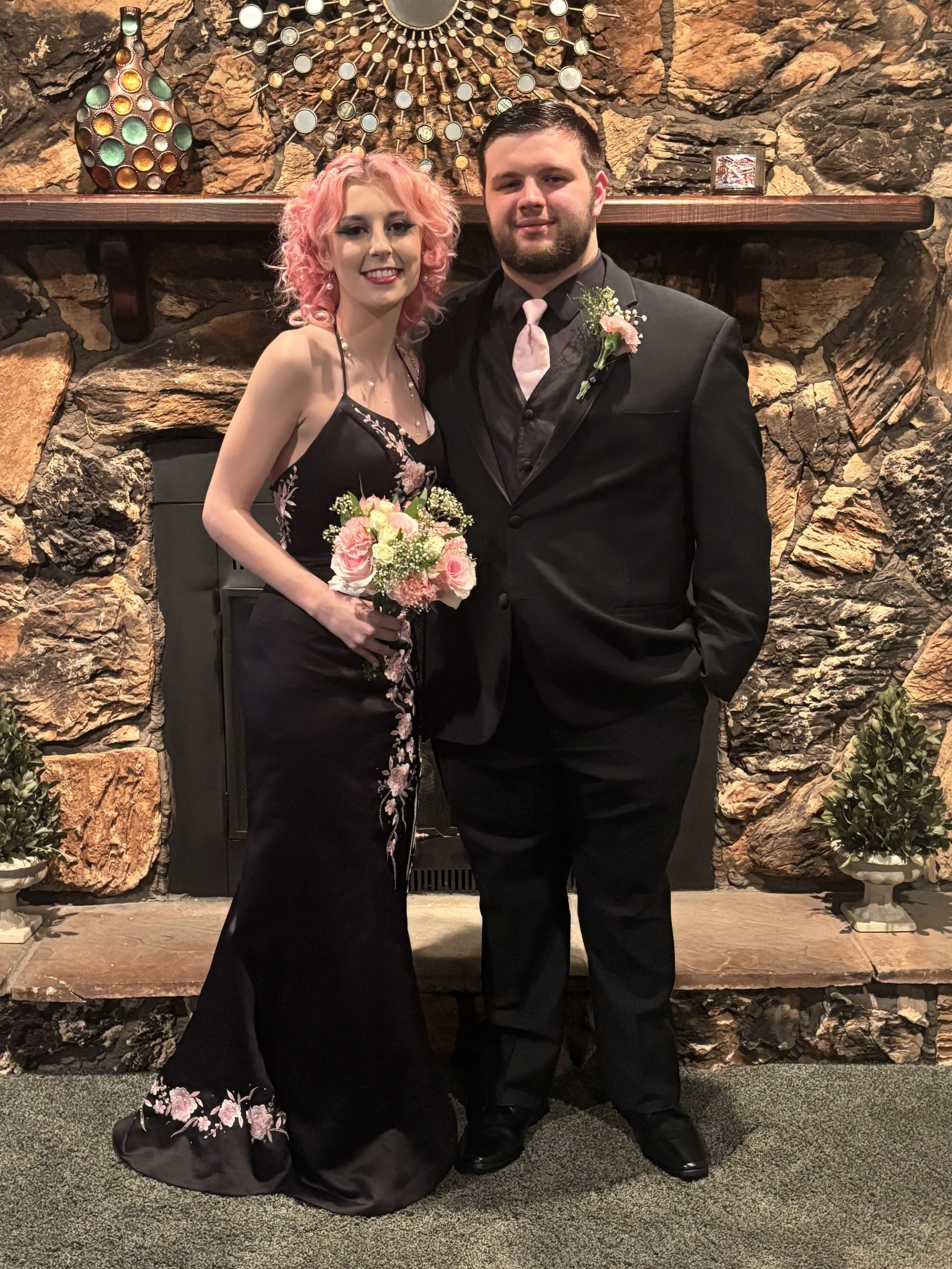 A young woman with pink curly hair and a man with short dark hair and a beard dressed in formal attire stand together, smiling, in front of a stone fireplace with decorative wall art and potted plants, holding a bouquet of pink and white flowers.