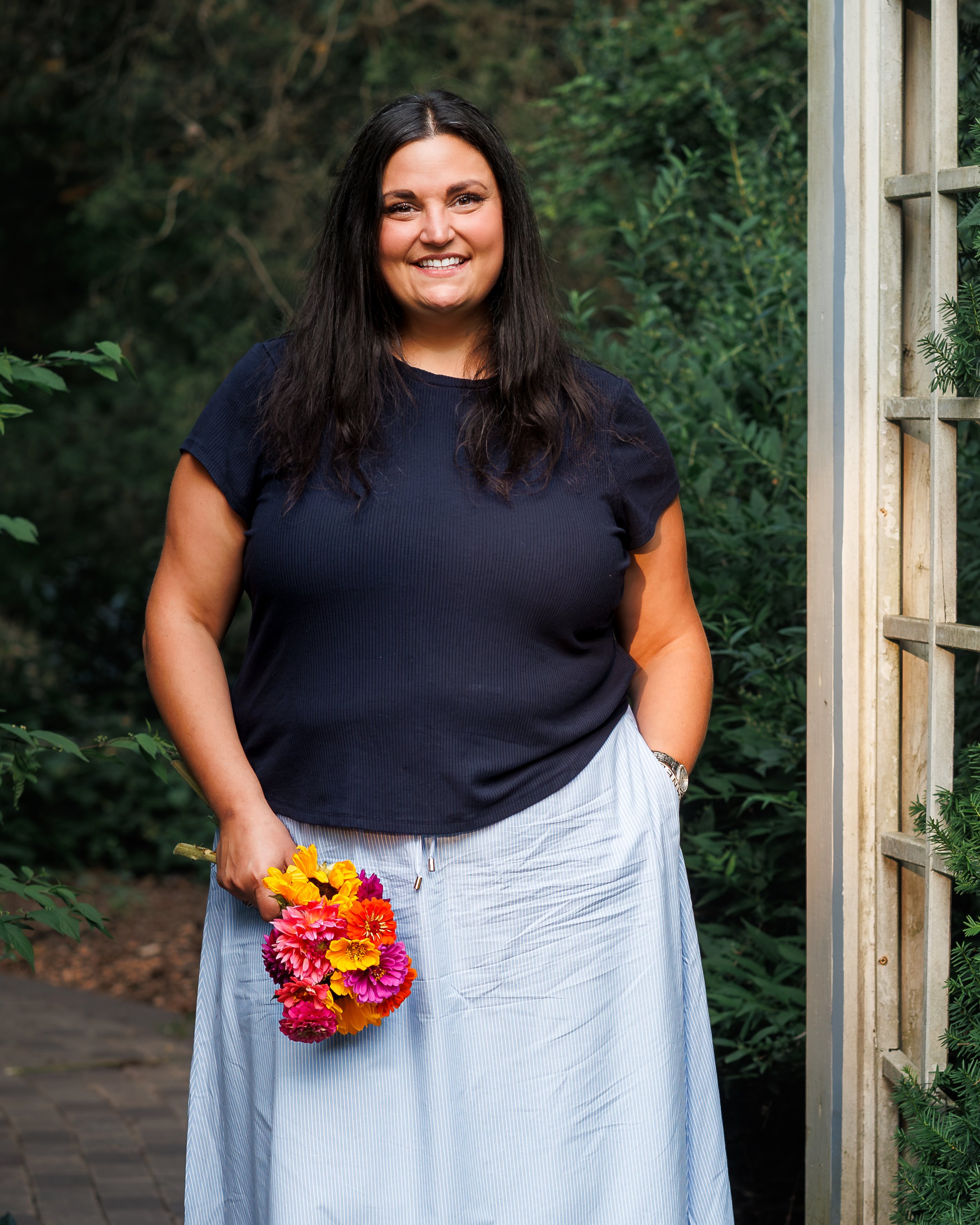 A woman with long dark hair and a bright smile holding a colorful bouquet of flowers, standing outdoors near a wooden fence and green foliage.