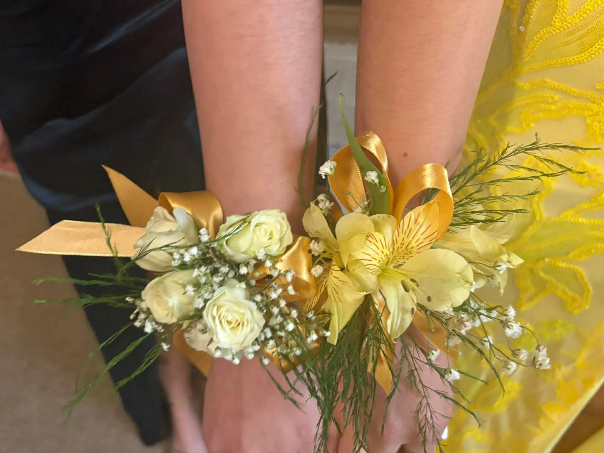 Close-up image of a bouquet of flowers with yellow lilies, white roses, small white filler flowers, and green foliage with a yellow satin ribbon, held in front of a person wearing a yellow dress and black clothing.