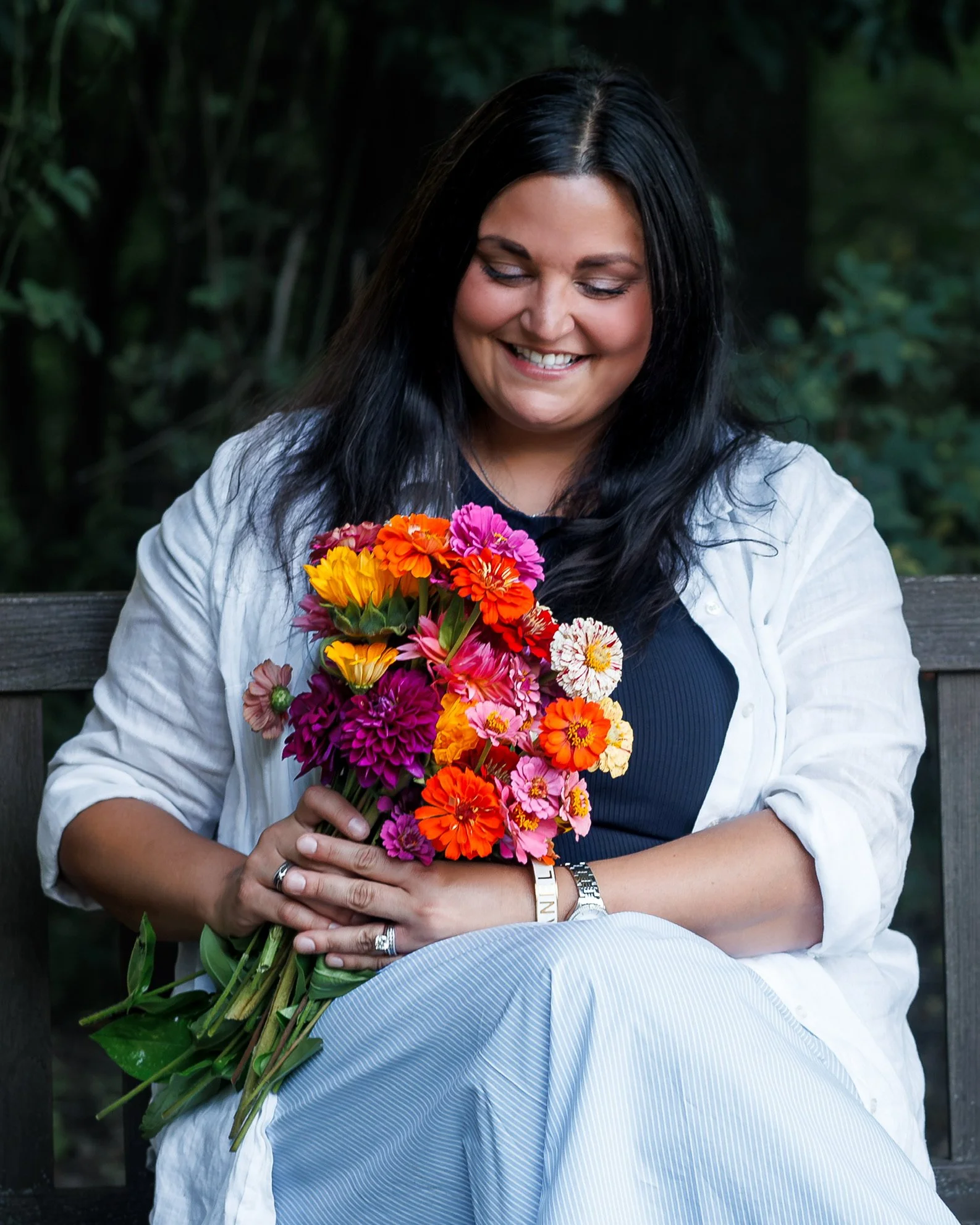 A woman with long dark hair, smiling and looking down, holding a large colorful bouquet of flowers including pink, orange, yellow, and purple blooms, sitting outdoors with a blurred green background.