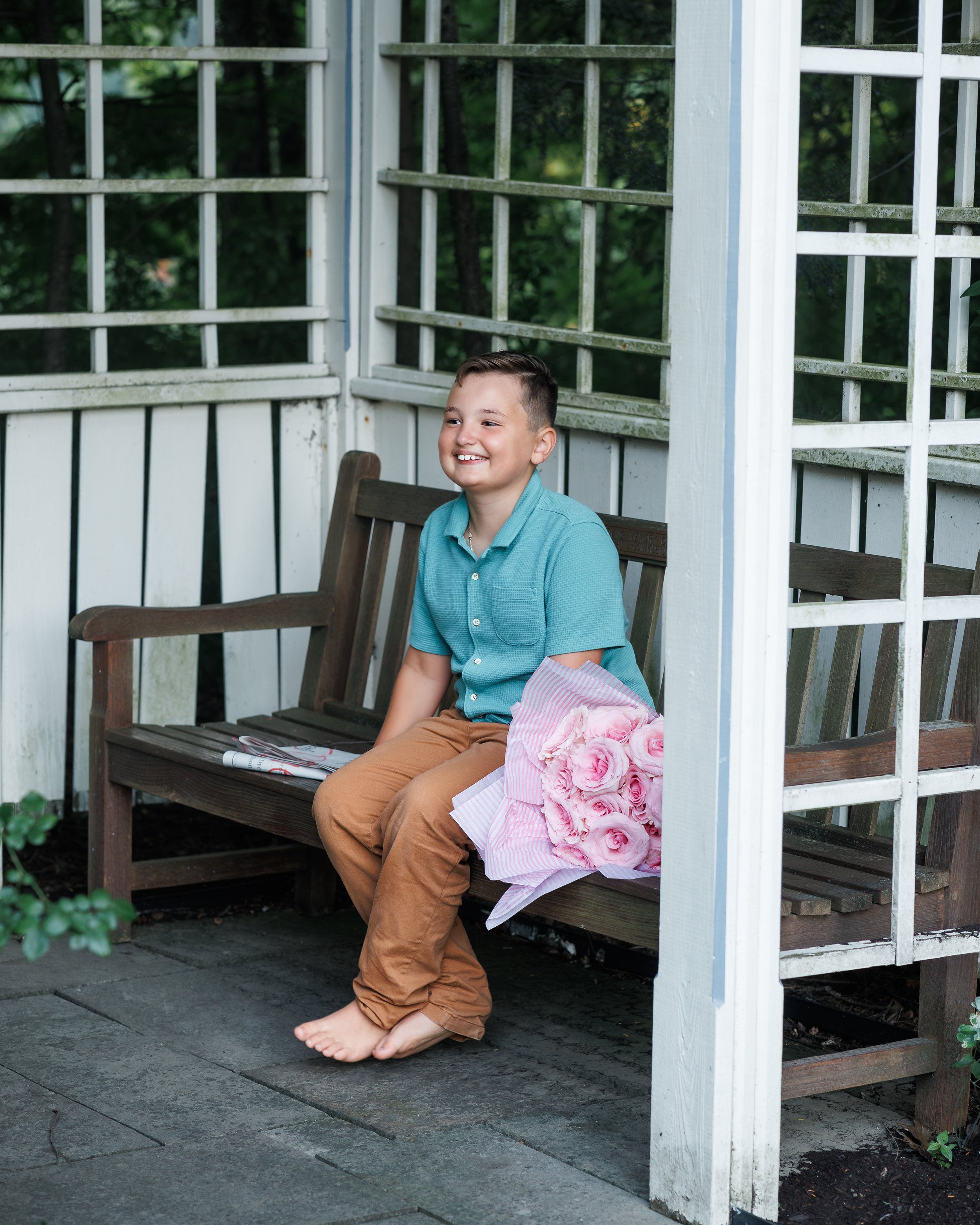 A young boy sitting barefoot on a wooden bench outdoors, smiling, with a bouquet of pink roses wrapped in pink paper resting on his lap, in front of a white lattice fence and green foliage.