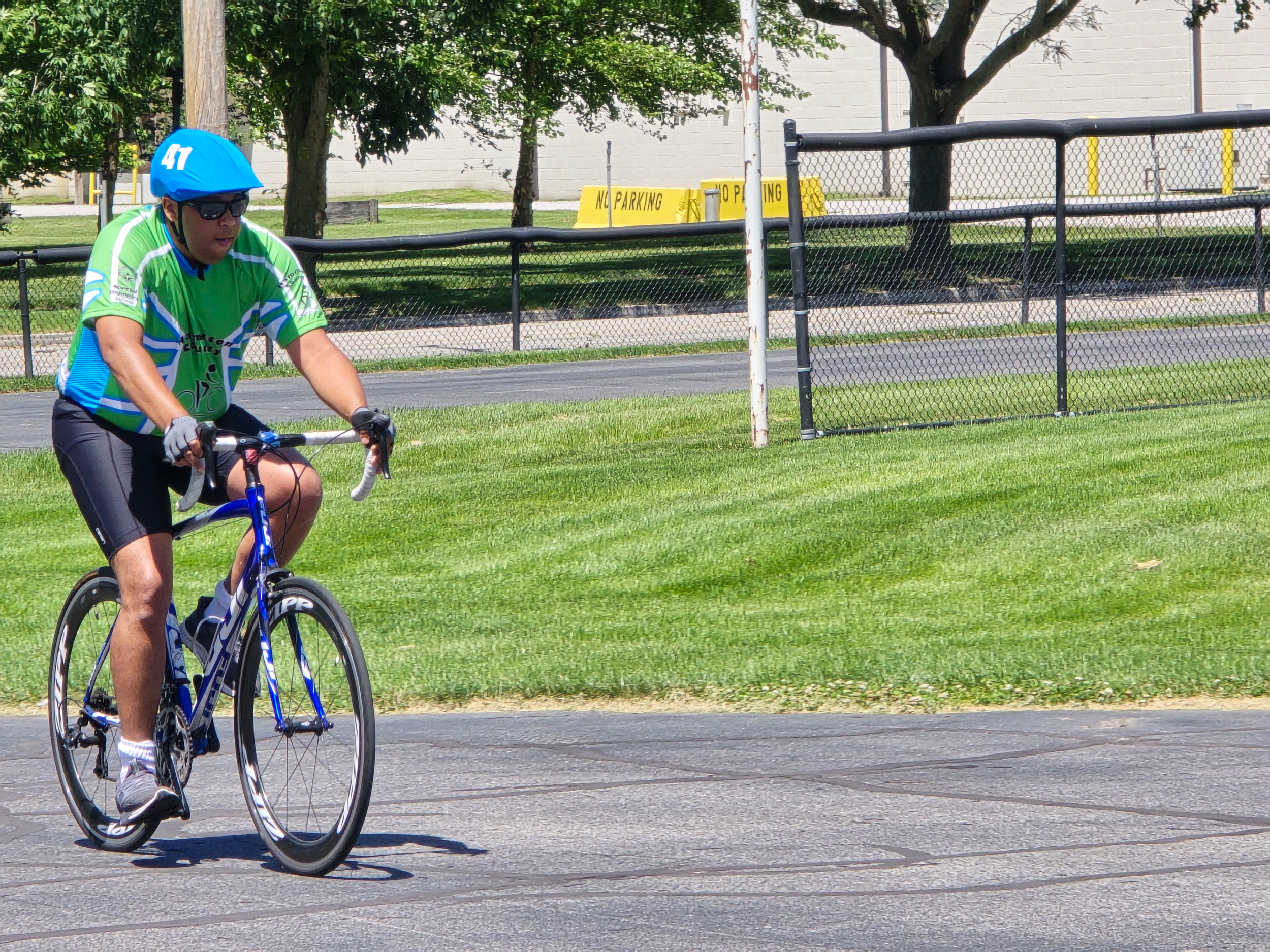 A man riding a blue bicycle on a paved path, wearing a bright green cycling jersey, black shorts, sunglasses, and a blue helmet with the number 41. There is a chain-link fence with a yellow "No Parking" sign and trees in the background.