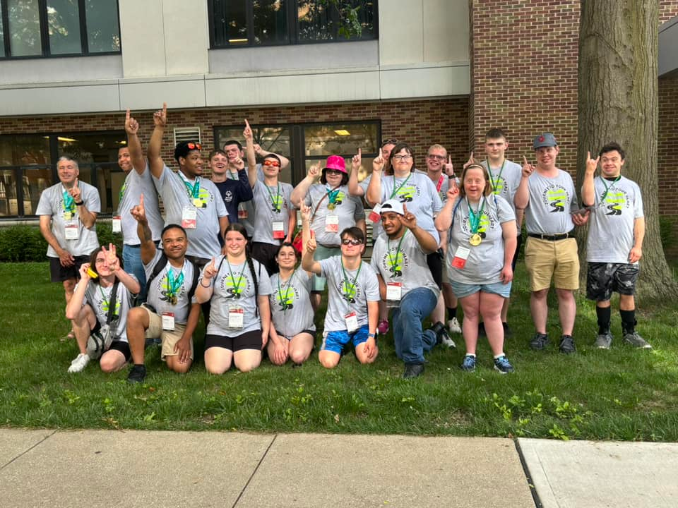 Group of young people and adults posing outdoors with some raising their hands and showing medals, in front of a brick building and tree.