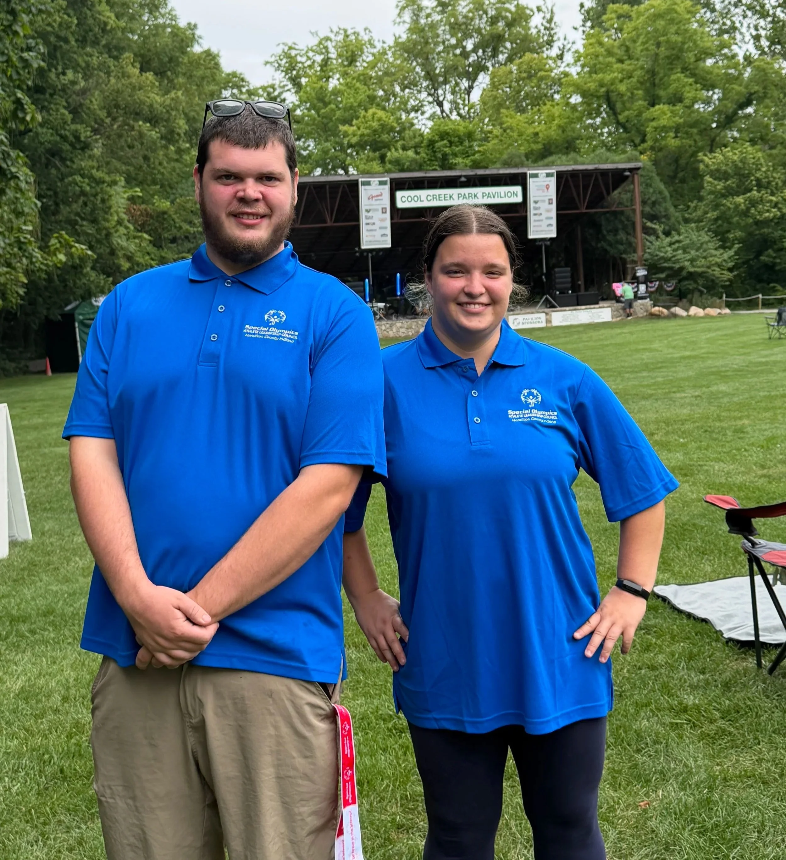 Two young individuals wearing blue polo shirts with the Special Olympics logo, standing outdoors on a grassy area with trees and a stage in the background. The stage has a sign that reads 'Cool Creek Park Pavilion.'