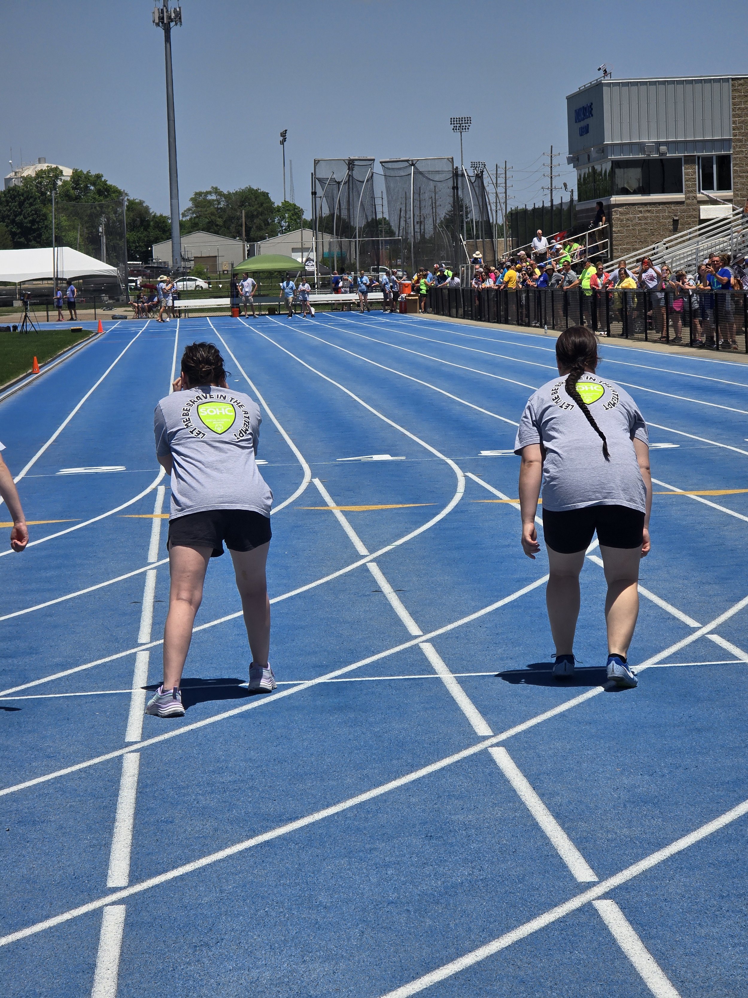 Two female athletes with braided hair wearing gray shirts and black shorts, preparing for a race on a blue running track outdoors, with spectators and officials in the background.