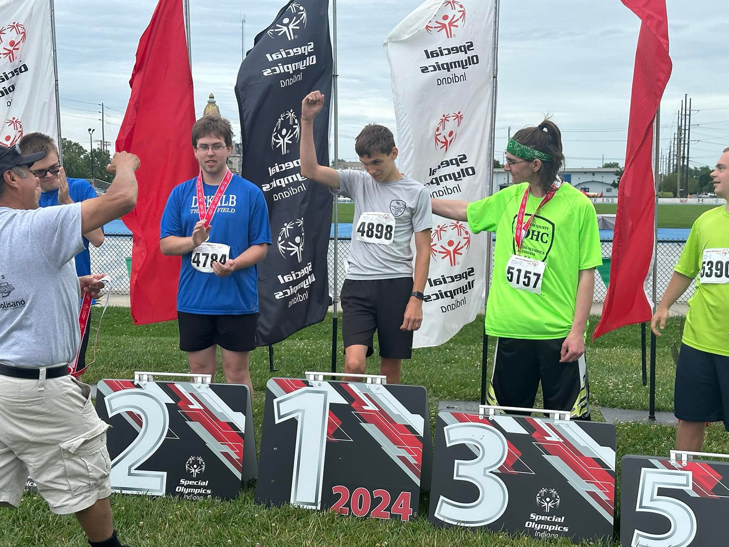 Young male athletes standing on a winners' podium at a Special Olympics event, surrounded by flags and banners, receiving medals. The first-place winner is raising his hand, and others hold medals and stand near their placement signs.
