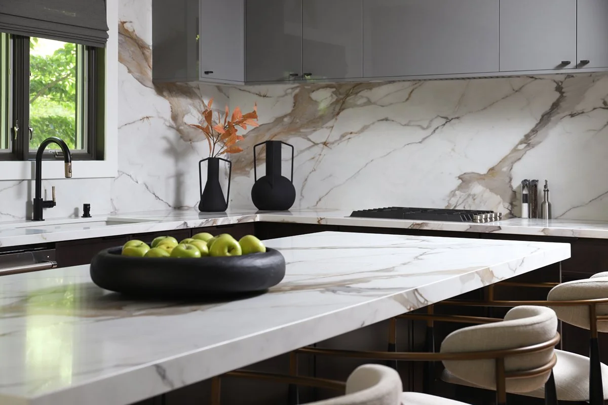 Modern kitchen with marble countertops and backsplash, black faucet, window revealing green foliage, decorative black vases with orange leaves, bowl of green apples, and cream-colored chairs around a marble island.