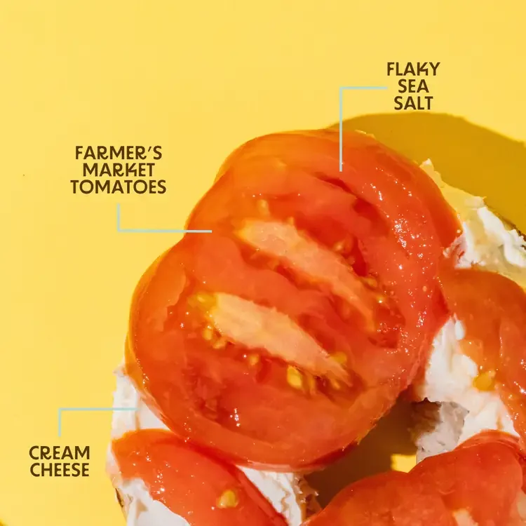 A tomato slice topped with flaky sea salt, served on cream cheese with a yellow background.