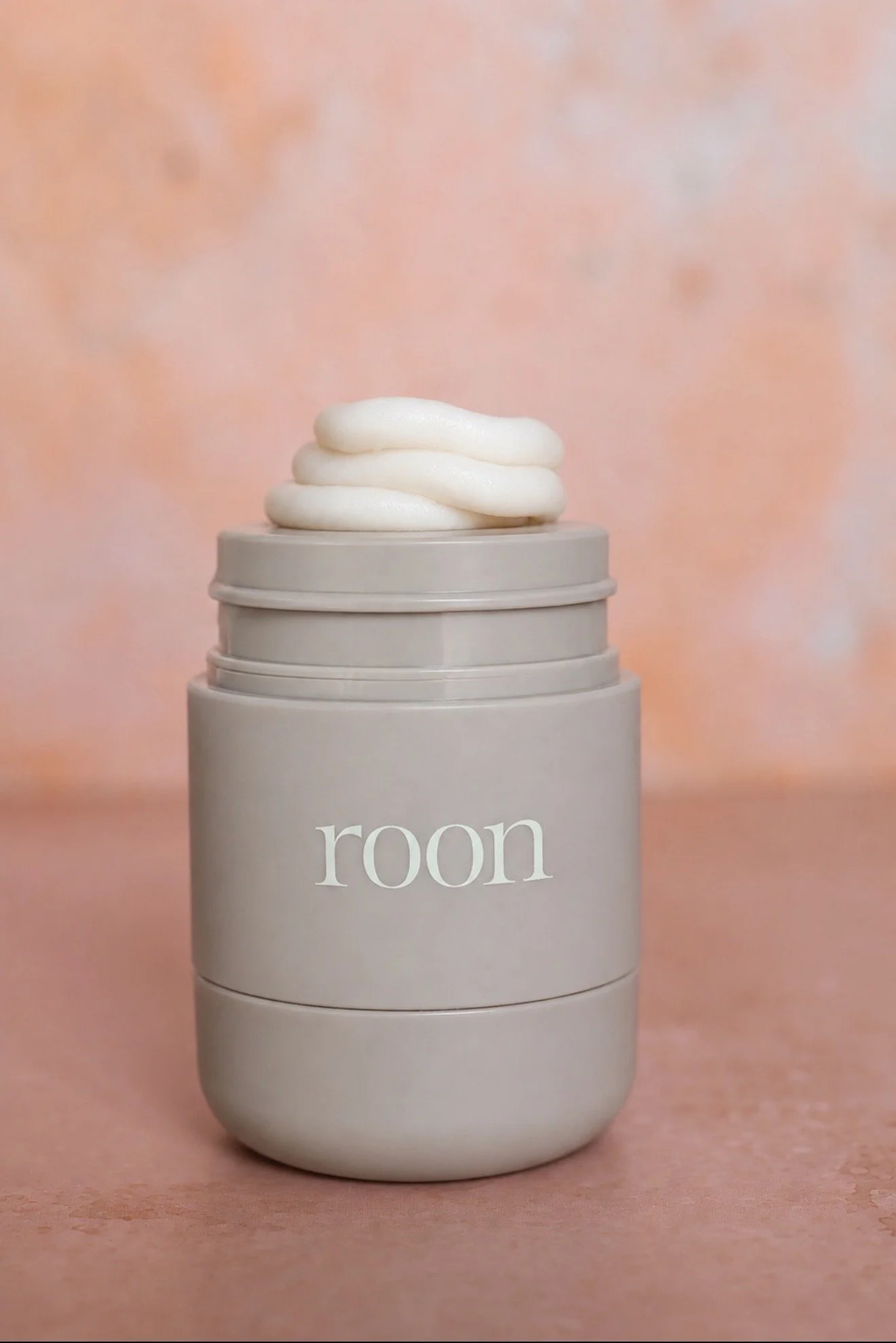 A cylindrical jar with a minimalist design, light gray color, and the word 'room' written on it in white letters. On top of the jar, there is a swirl of white cream or foamy substance, set against a pinkish-orange background.