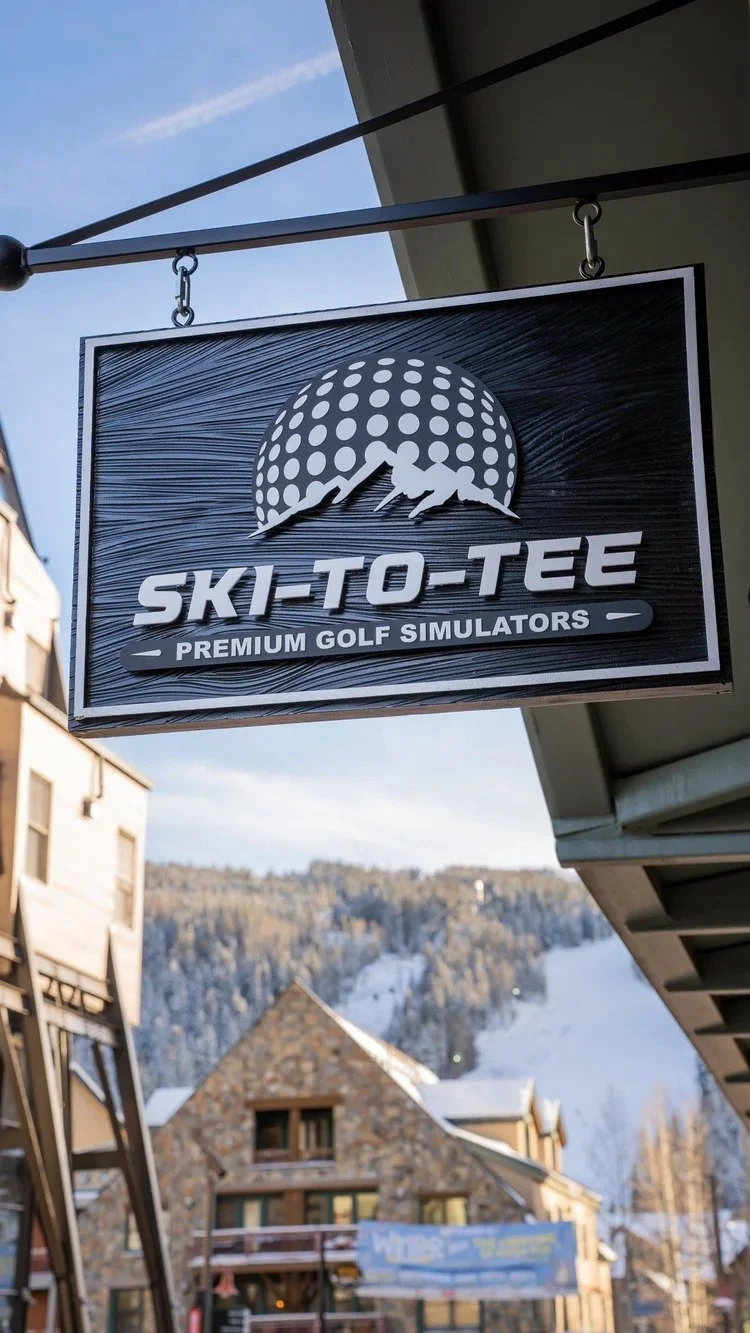 A rectangular sign hanging outside a building reads 'SKI-TO-TEE PREMIUM GOLF SIMULATORS' with a logo of a golf ball and two mountaineers climbing a mountain in the background. The sky is clear and snowy hills and a stone building are visible in the d