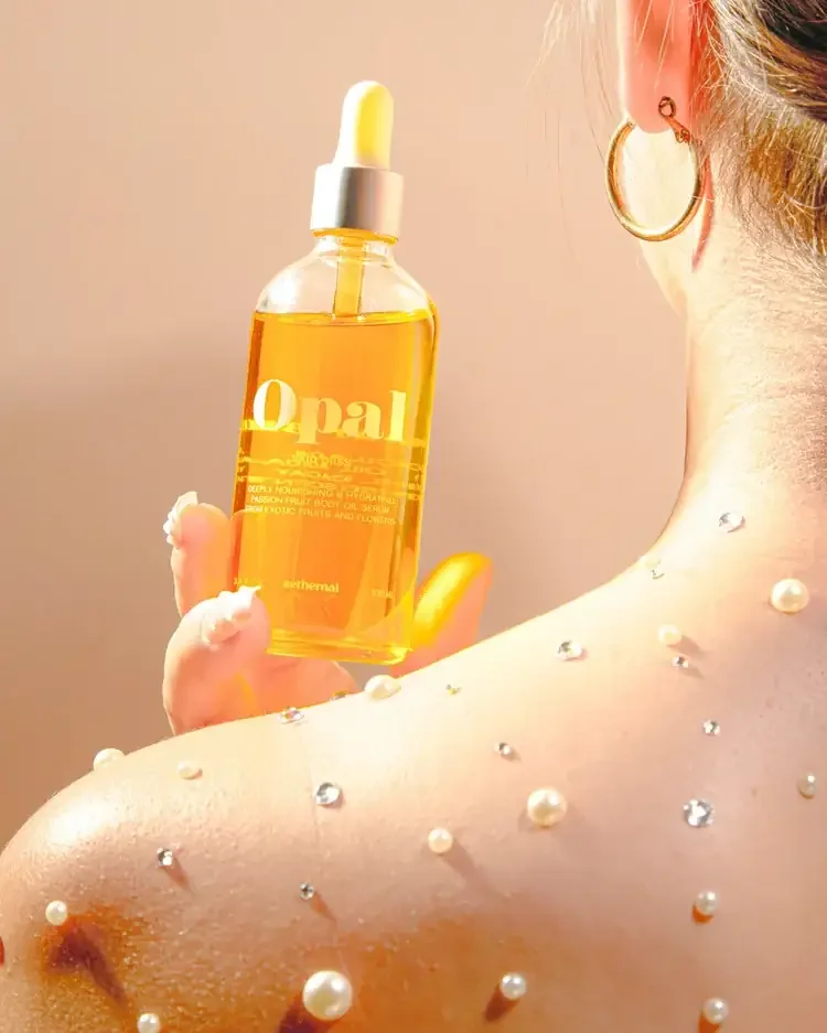 Close-up of a woman holding a yellow serum bottle with a dropper, earrings on her ear, and beads or pearls on her shoulder, against a neutral background.