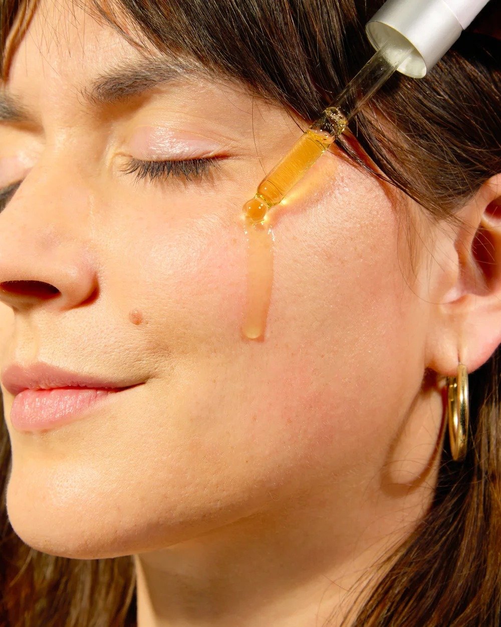 A woman receiving a facial treatment with a dropper applying a golden-colored serum to her cheek.